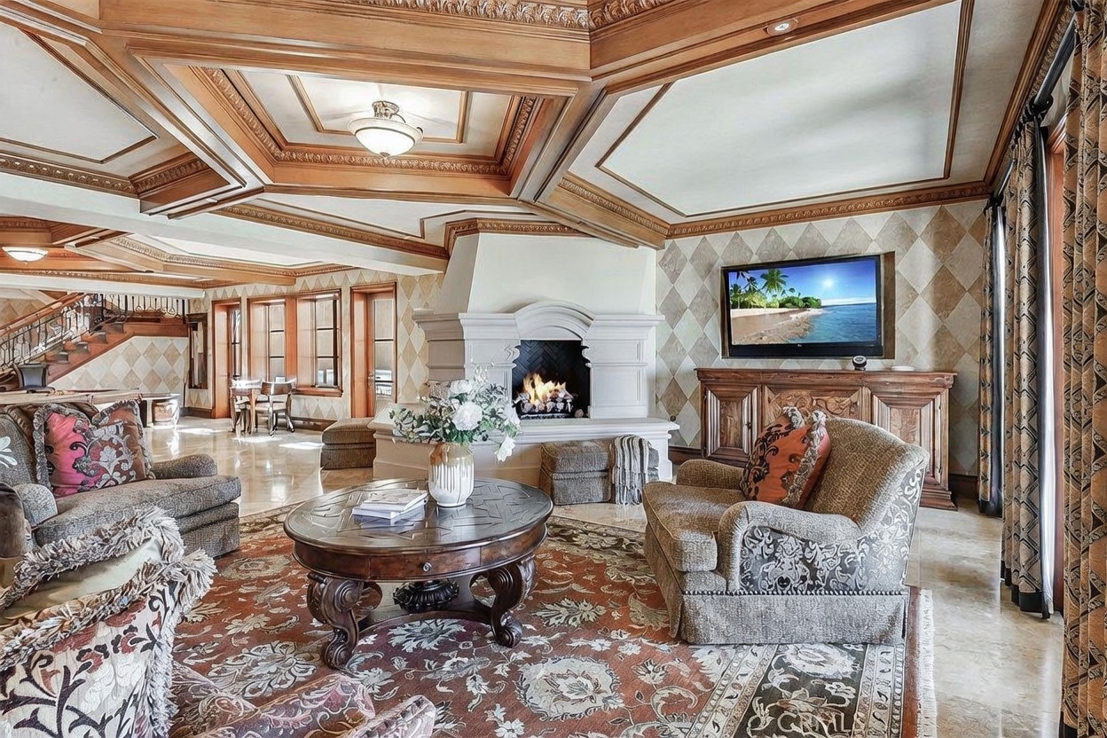 This spacious living room features an ornate coffered wood ceiling and a prominent stone fireplace with a decorative mantel. The room is furnished with plush patterned armchairs and a large circular coffee table set atop a detailed area rug. A television is mounted on the wall above a wooden media console, and the space is accented by diamond-patterned wallpaper and warm, ambient lighting.