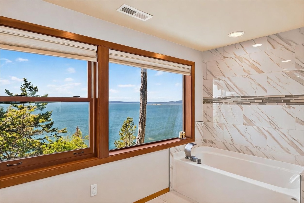 This primary bathroom features a deep soaking tub positioned directly beneath large windows that offer a panoramic view of the ocean and distant coastline. The walls are finished with light-toned, veined stone tiles accented by a horizontal decorative mosaic strip. The space is bright and airy, emphasizing a serene, spa-like atmosphere with a focus on the natural scenery outside.