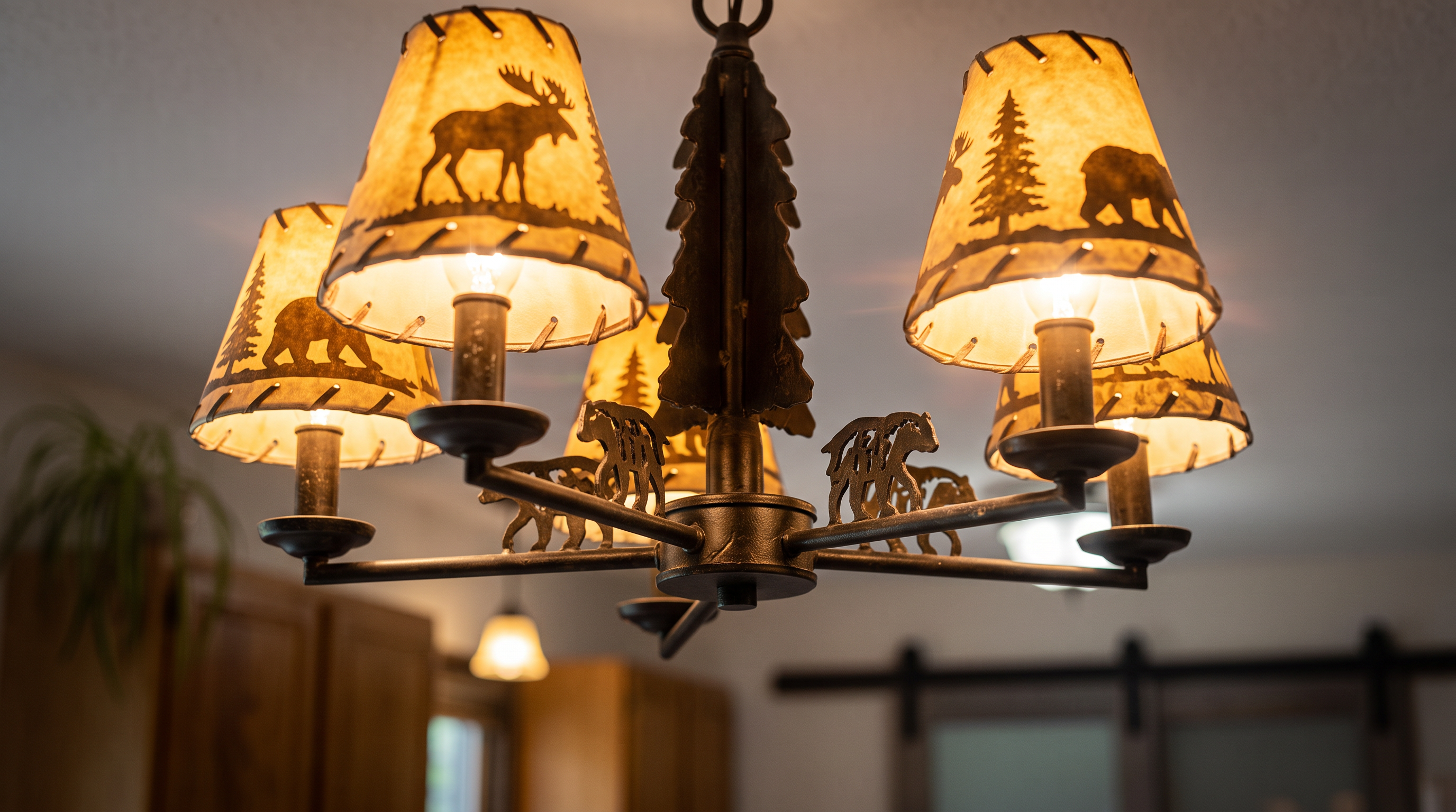 This image features a rustic-style chandelier characterized by a lodge-inspired design. The fixture includes metal accents shaped like pine trees and wildlife silhouettes, paired with parchment-style shades depicting deer and moose forest scenes, which provide warm ambient lighting.