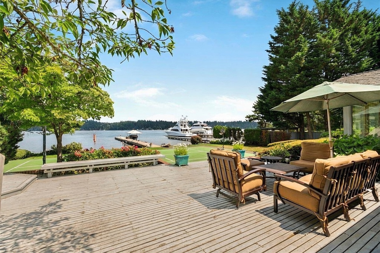 This expansive wooden deck offers a serene outdoor living space overlooking a waterfront view with private boat docks. The deck is furnished with comfortable patio seating under a large umbrella, providing a perfect vantage point to enjoy the lush green trees and the calm water beyond. The perspective captures the transition from the private deck to the manicured lawn and the scenic harbor, creating an inviting and tranquil atmosphere.