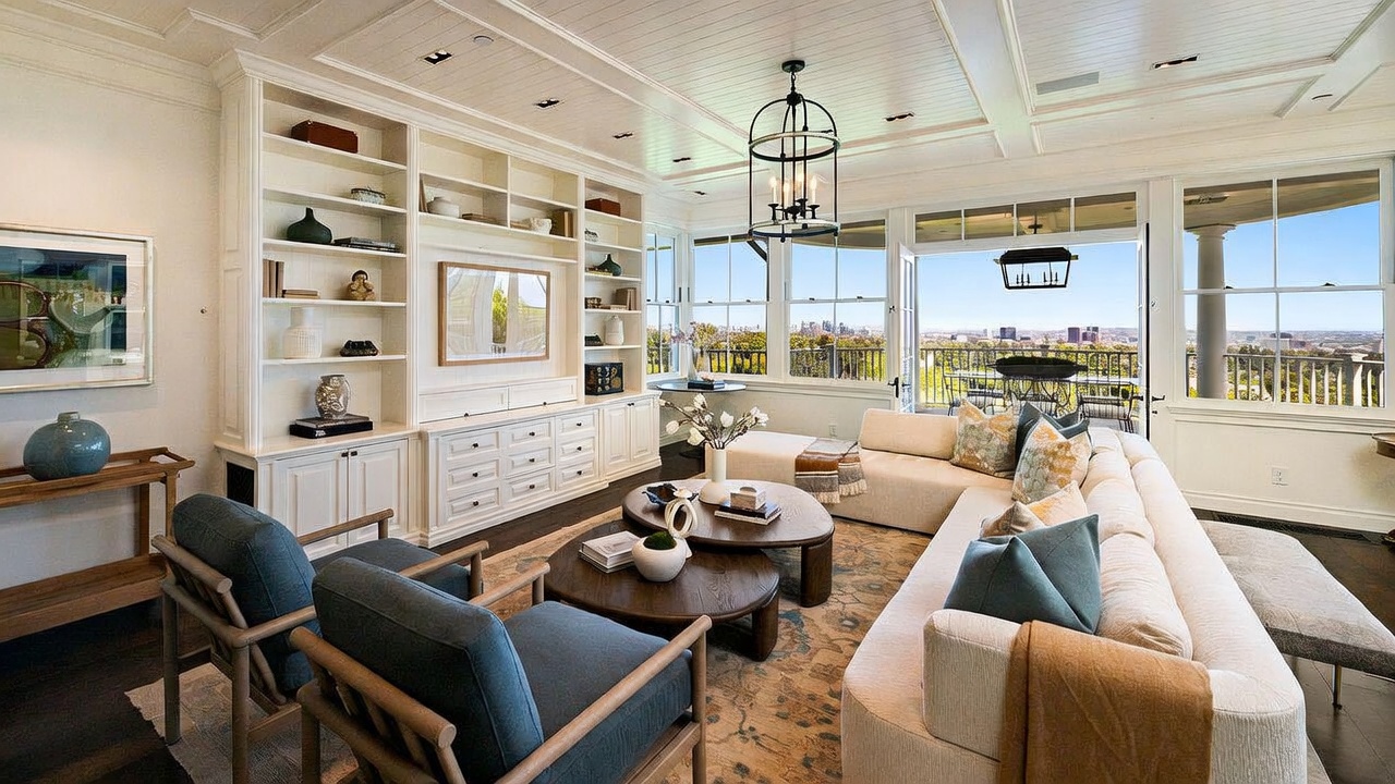 This bright and airy living room features a large, neutral-toned sectional sofa and two blue armchairs arranged around a pair of circular wooden coffee tables. The space is anchored by a built-in white shelving unit and cabinetry that spans the wall, while large windows and glass doors provide an expansive view of the city skyline. A coffered ceiling with a decorative lantern-style chandelier adds a touch of elegance to this sophisticated, open-concept living area.