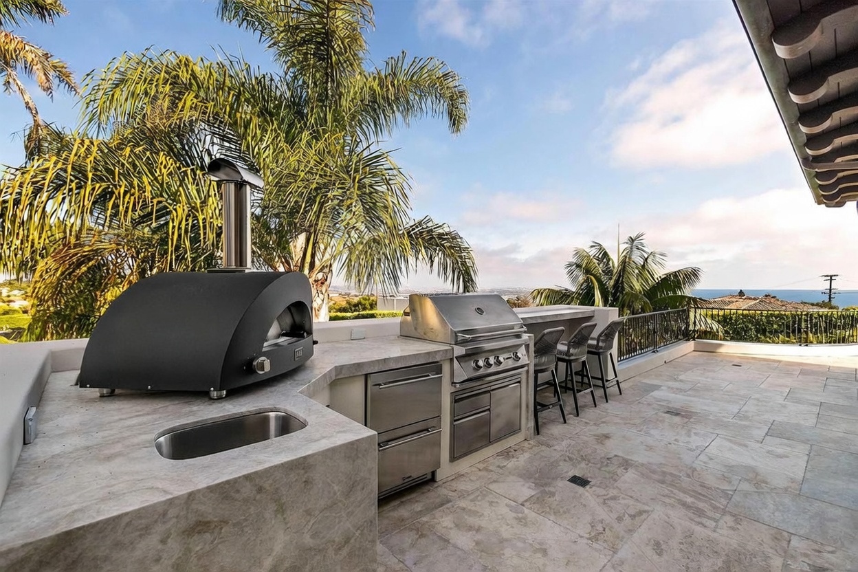 This expansive outdoor kitchen and patio area features a stone-topped counter equipped with a stainless steel grill, a pizza oven, and a built-in sink. The space includes a bar-style seating arrangement with three stools, overlooking a scenic coastal view with palm trees in the background. The stone-tiled flooring and open-air design create a luxurious, resort-like atmosphere perfect for outdoor entertaining.