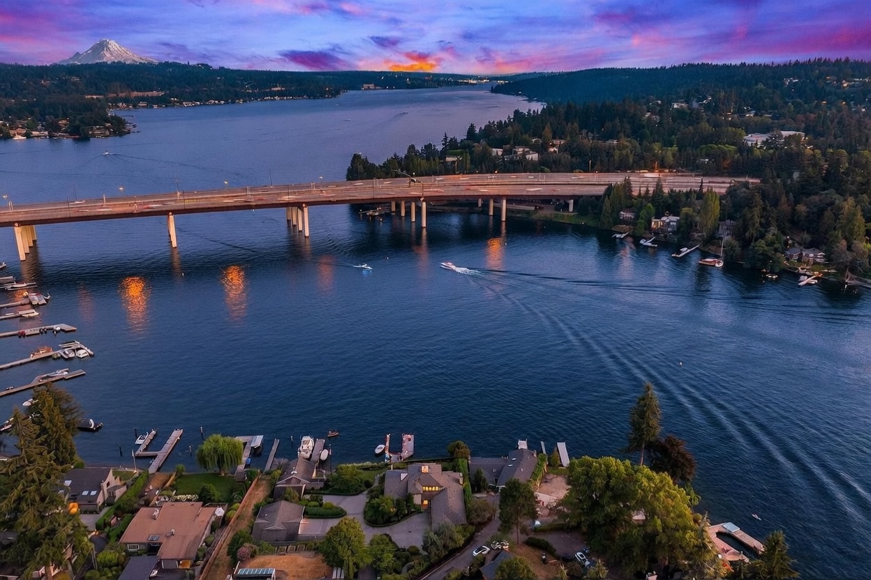 This stunning aerial perspective captures a waterfront property at dusk, showcasing its prime location along a serene lake with a prominent bridge spanning the water in the background. The scene features a lush, landscaped estate with a private dock, mature trees, and a multi-level residence, all set against a dramatic sunset sky with a distant mountain peak. The composition emphasizes the expansive water views and the tranquil, high-end lifestyle associated with this prime real estate.