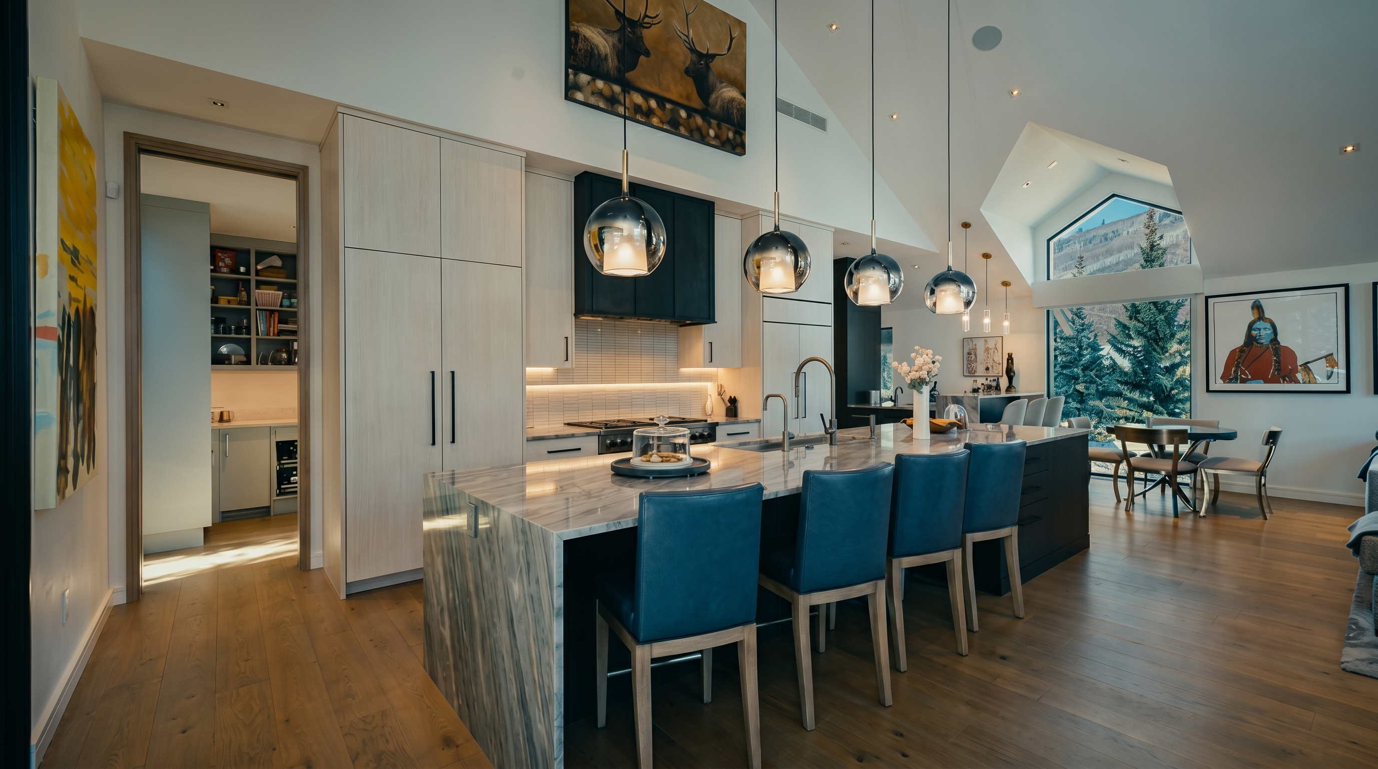 This spacious, open-concept kitchen features a large central island with a stone countertop and four blue leather-upholstered stools. The area is illuminated by four modern glass pendant lights hanging from a vaulted ceiling, with light wood flooring extending throughout the space. A doorway leads to a secondary pantry area, while large windows in the background provide a scenic view of a snowy, wooded landscape.