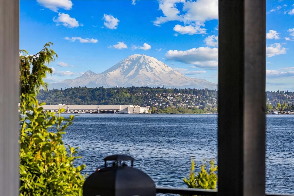 This view is captured from inside a room, looking out through a large window frame that acts as a natural border for the scenery. The primary focus is a breathtaking, unobstructed view of a snow-capped mountain rising above a body of water and a distant shoreline with residential homes. In the foreground, a portion of a dark lantern and some green foliage are visible, adding depth and a sense of being indoors looking out at the landscape.