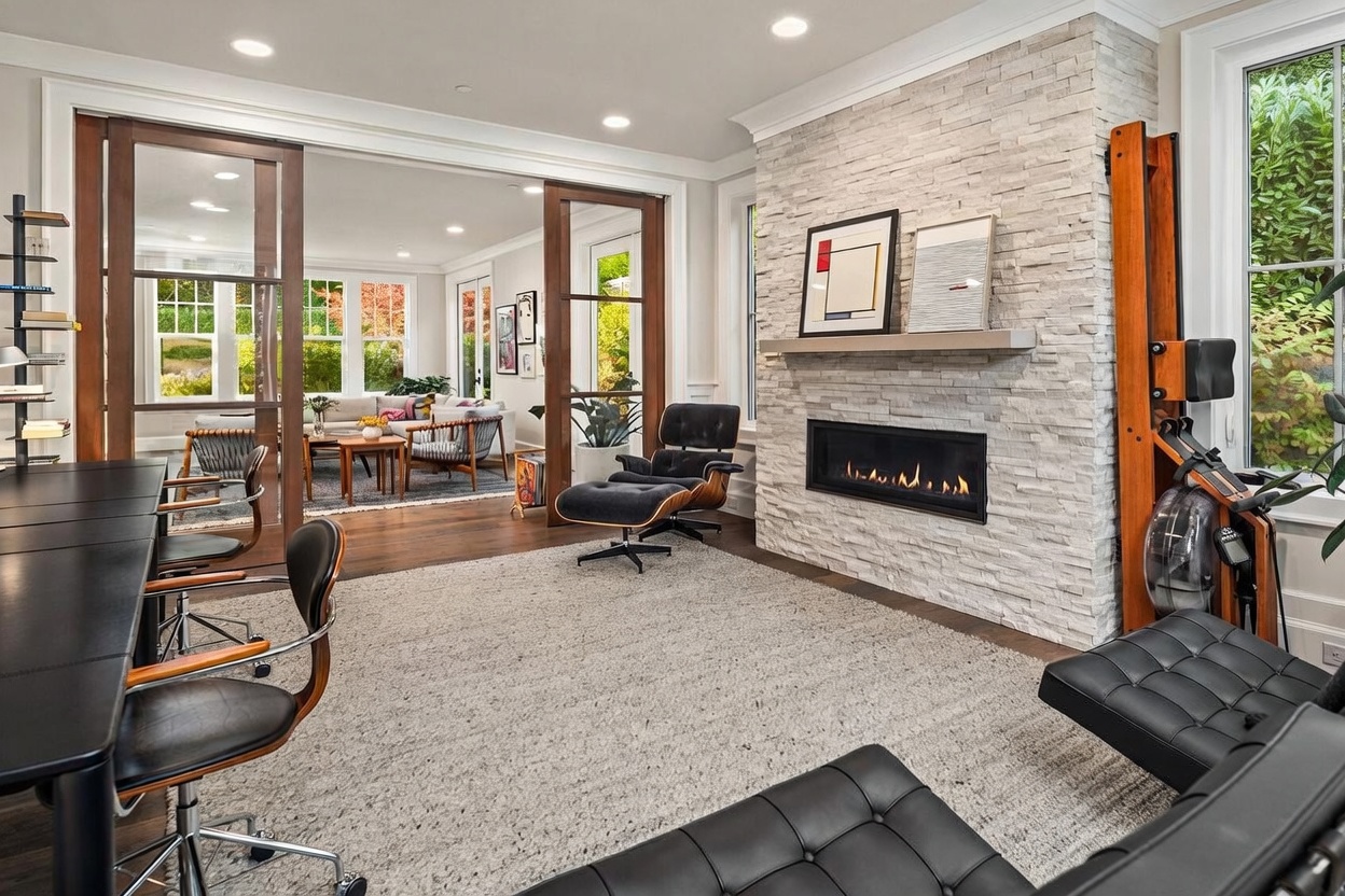 This sophisticated home office features a sleek black desk, a classic lounge chair with an ottoman, and a prominent stacked-stone chimney breast housing a linear fireplace. Large sliding glass doors with wood frames provide a seamless transition to an adjacent living area, creating an open and airy atmosphere. The room is accented by a textured area rug, a rowing machine, and modern art, blending professional utility with comfortable, high-end living.