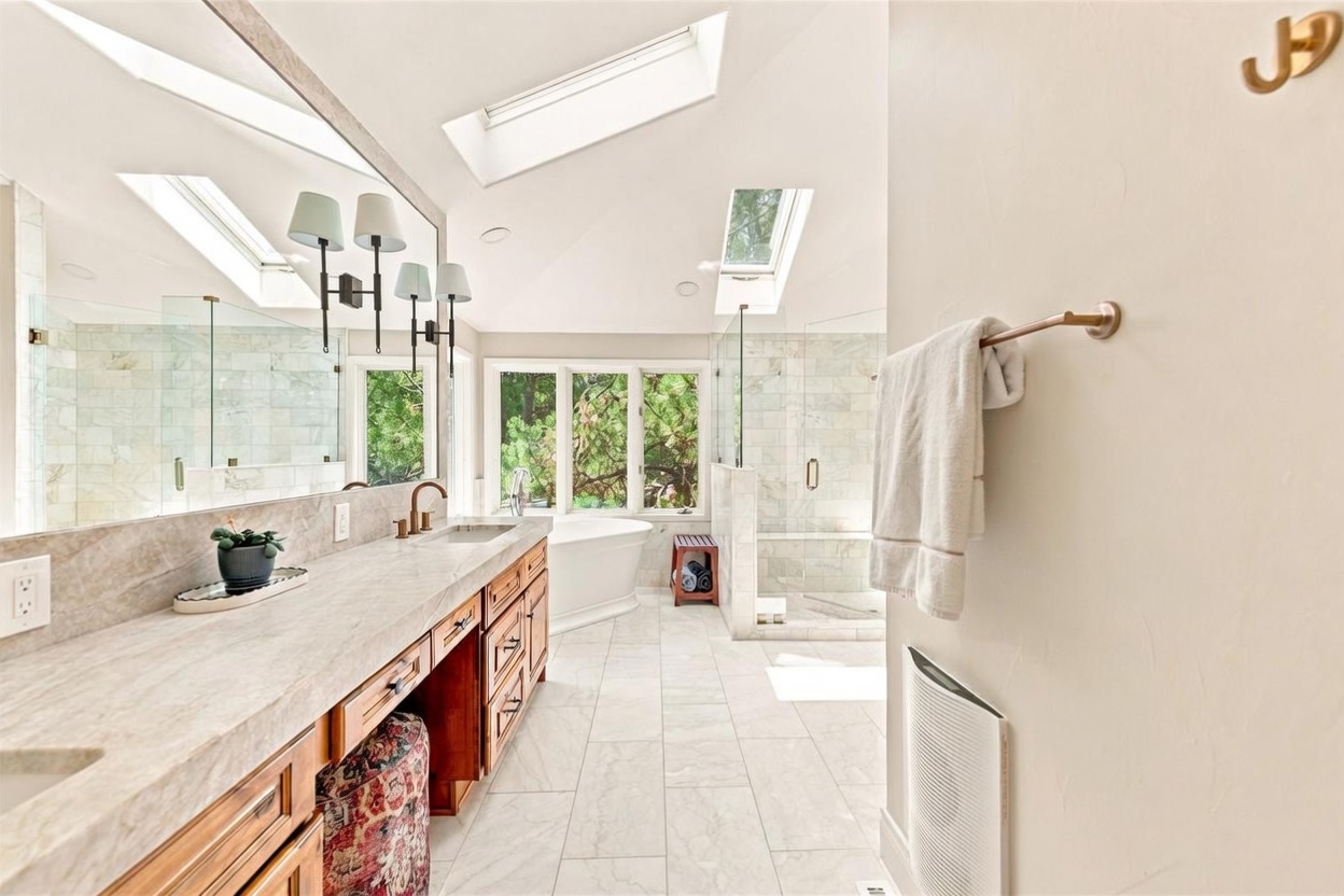 This spacious primary bathroom features a double vanity with stone countertops, warm wood cabinetry, and a large mirror illuminated by wall-mounted light fixtures. A freestanding soaking tub sits beneath a window, while a separate glass-enclosed shower with stone tile walls is positioned to the right. The room is bright and airy, enhanced by multiple skylights that flood the space with natural light.
