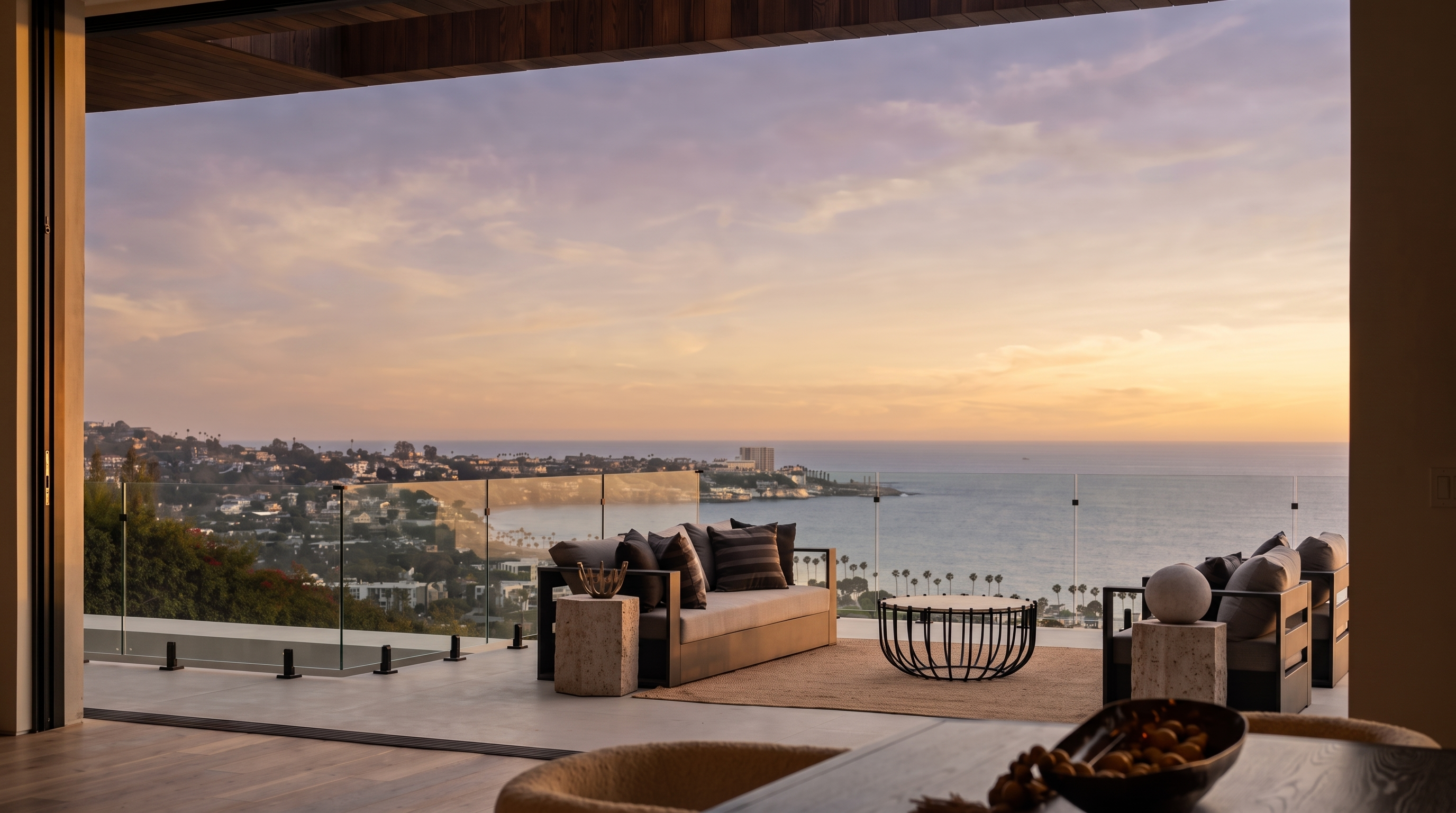 This expansive outdoor terrace offers a seamless transition from the interior living space, featuring a modern seating area with a sofa and a circular coffee table. The balcony is enclosed by a sleek glass railing that provides an unobstructed, panoramic view of the coastline, ocean, and sunset-lit sky. The perspective is captured from inside looking out, emphasizing the luxurious indoor-outdoor lifestyle and the dramatic coastal scenery.