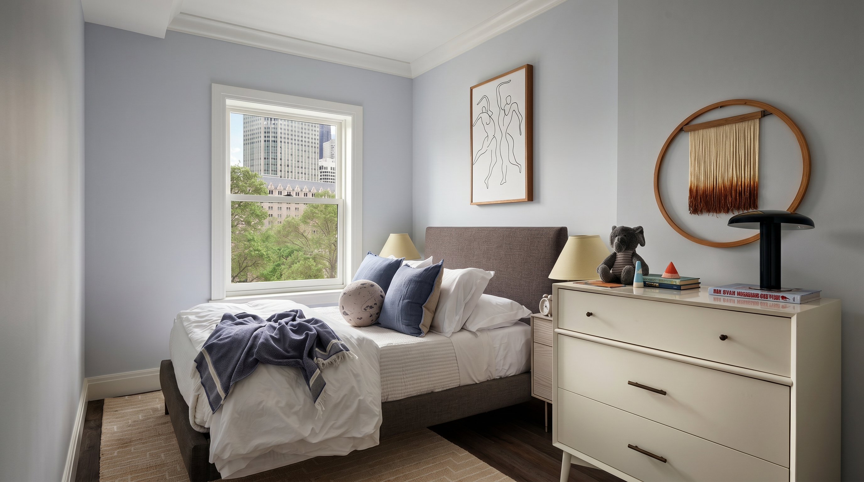 This cozy, well-lit bedroom features a dark upholstered bed frame contrasted against soft light blue walls and dark hardwood flooring. A large window provides a view of urban greenery and city buildings, while a white mid-century modern dresser and minimalist wall art add a clean, contemporary aesthetic. The room is accented by a textured area rug and a circular wall hanging, creating a warm and inviting atmosphere.