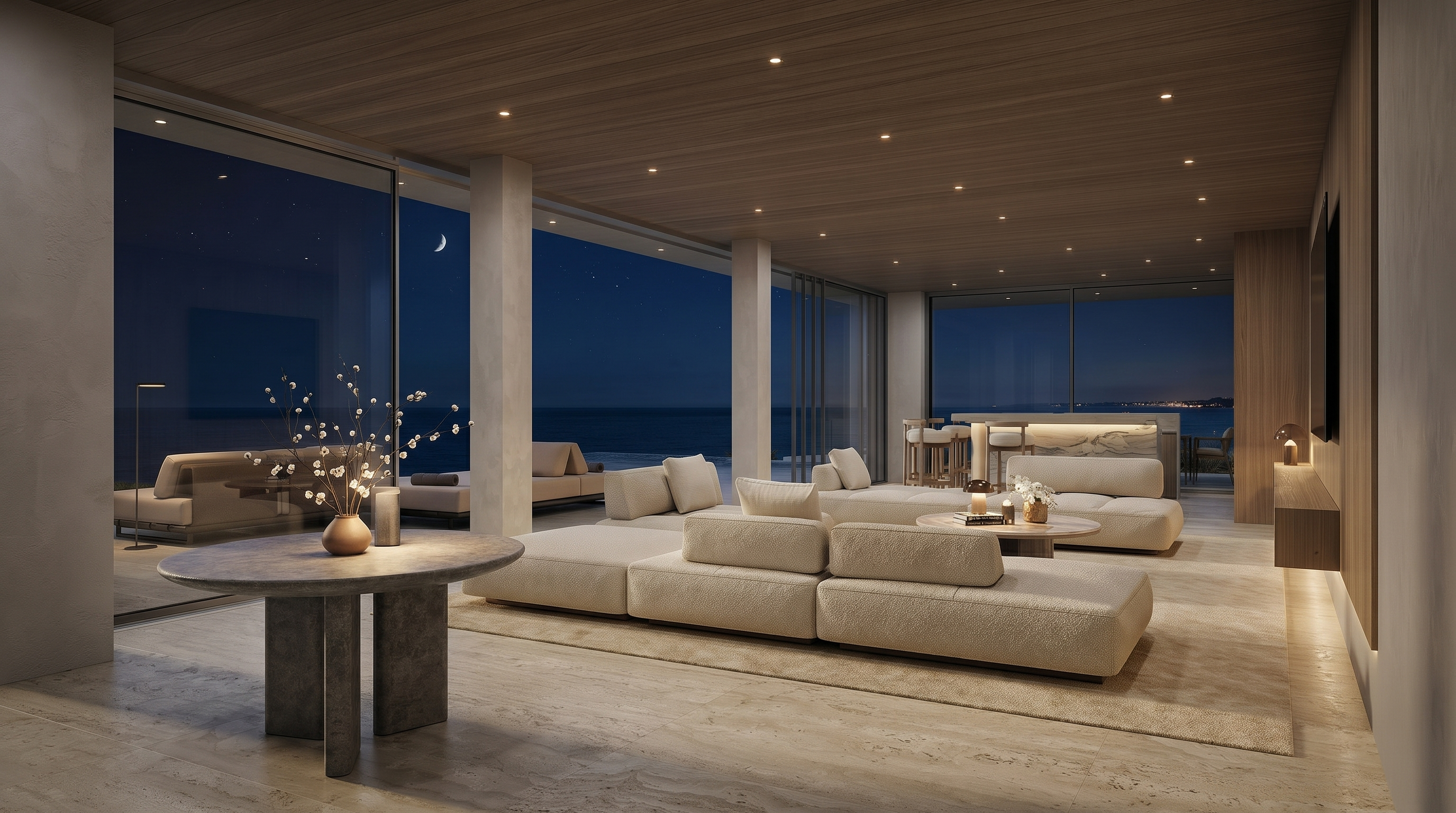 This expansive, open-concept living area features a neutral color palette and a seamless transition to the outdoors through floor-to-ceiling glass walls. The space is furnished with plush, low-profile modular seating arranged on a large area rug, complemented by a circular stone table in the foreground. The room is defined by a light wood-paneled ceiling with recessed lighting and a stunning, unobstructed view of the ocean at sunset.