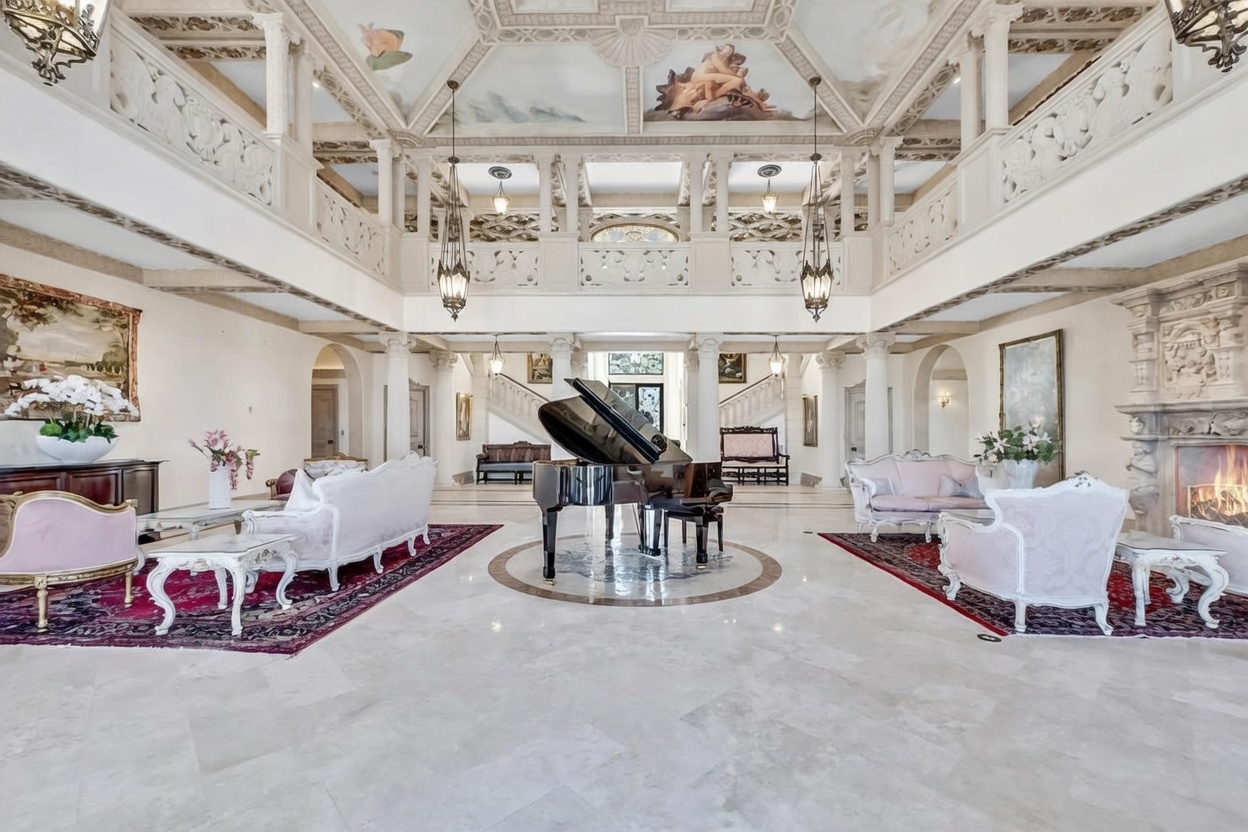 This grand, double-height living room features an opulent, neoclassical design with a painted ceiling mural and ornate architectural detailing. A polished stone floor reflects the light from two large chandeliers, while a central grand piano serves as a focal point between two symmetrical seating areas. The space is framed by white columns and a balcony overlook, creating an expansive and luxurious atmosphere.