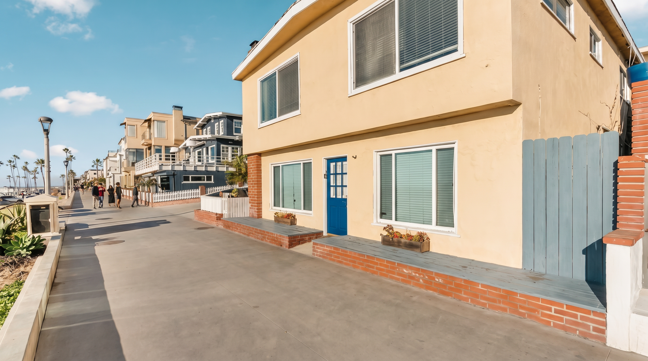 This exterior view captures a two-story, light-colored stucco home situated directly along a paved beach boardwalk. The house features a prominent blue front door, multiple rectangular windows with white frames, and a low brick planter box running along the base of the front facade. The perspective is from the boardwalk, showcasing the home's immediate proximity to the public path and the coastal environment.