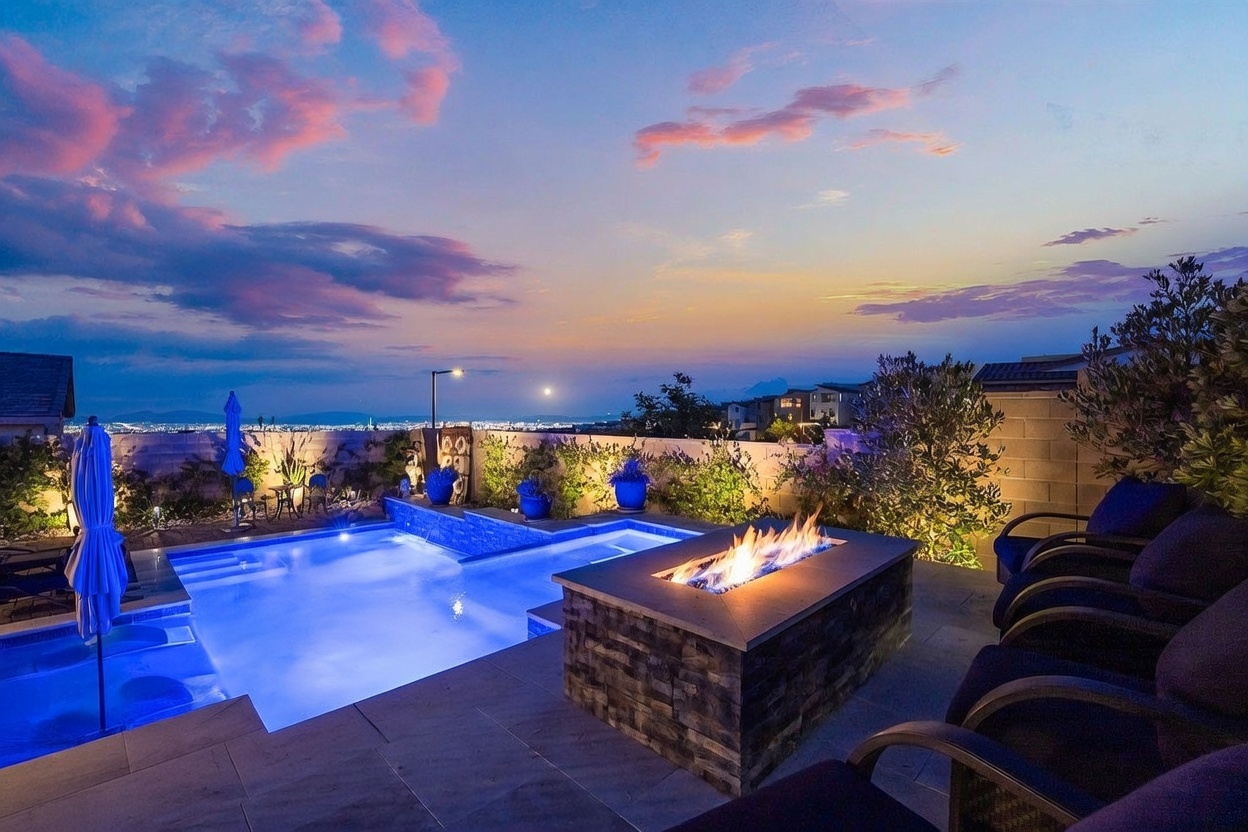 This stunning outdoor living space features a modern rectangular pool and integrated spa, set against a dramatic sunset sky overlooking a city view. A stone-clad fire pit serves as a central gathering point on the paved patio, surrounded by comfortable lounge seating. The area is framed by a decorative metal railing and lush landscaping, creating an inviting atmosphere for evening relaxation.