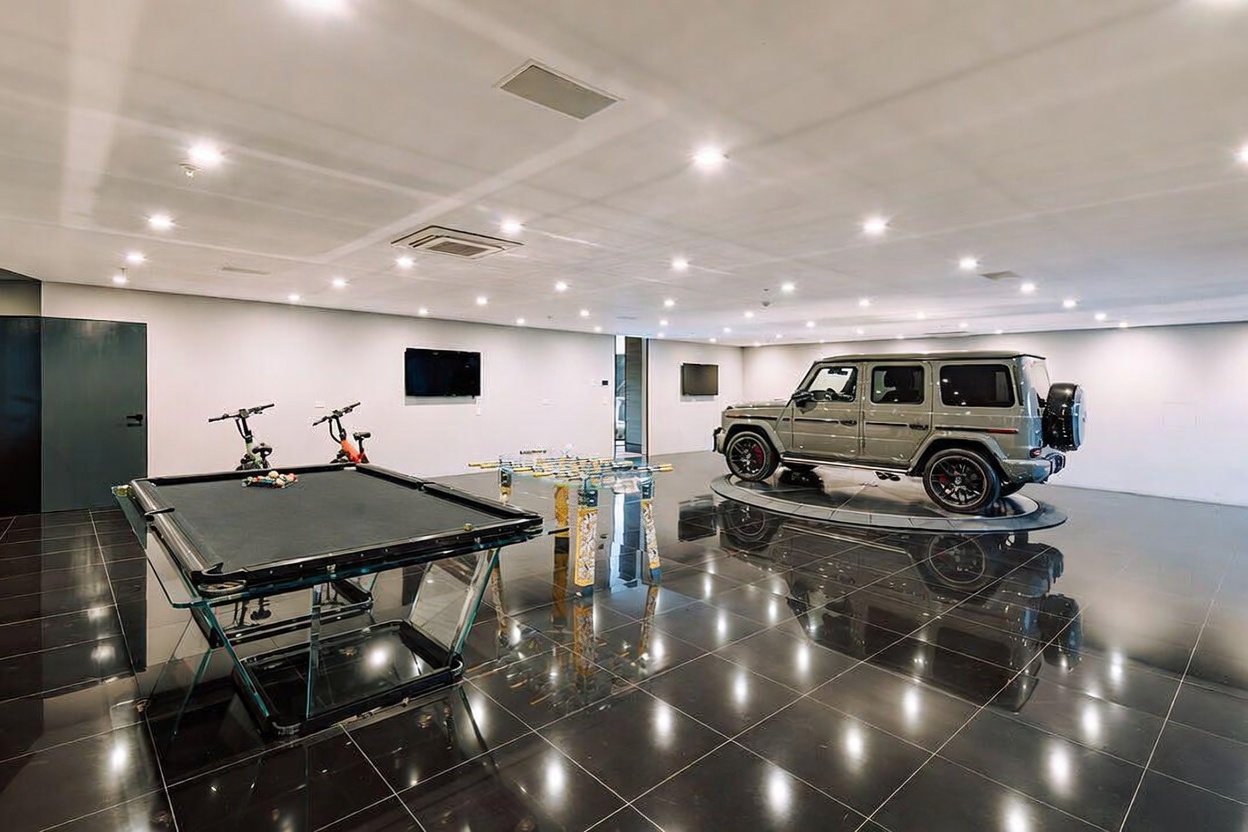 This expansive, modern garage space doubles as a luxury entertainment area, featuring a high-gloss black tiled floor and a rotating display platform for a premium SUV. The room is equipped with a glass-topped pool table and a foosball table, creating a sophisticated lounge atmosphere. Recessed ceiling lighting and minimalist white walls provide a clean, gallery-like backdrop for the vehicle.
