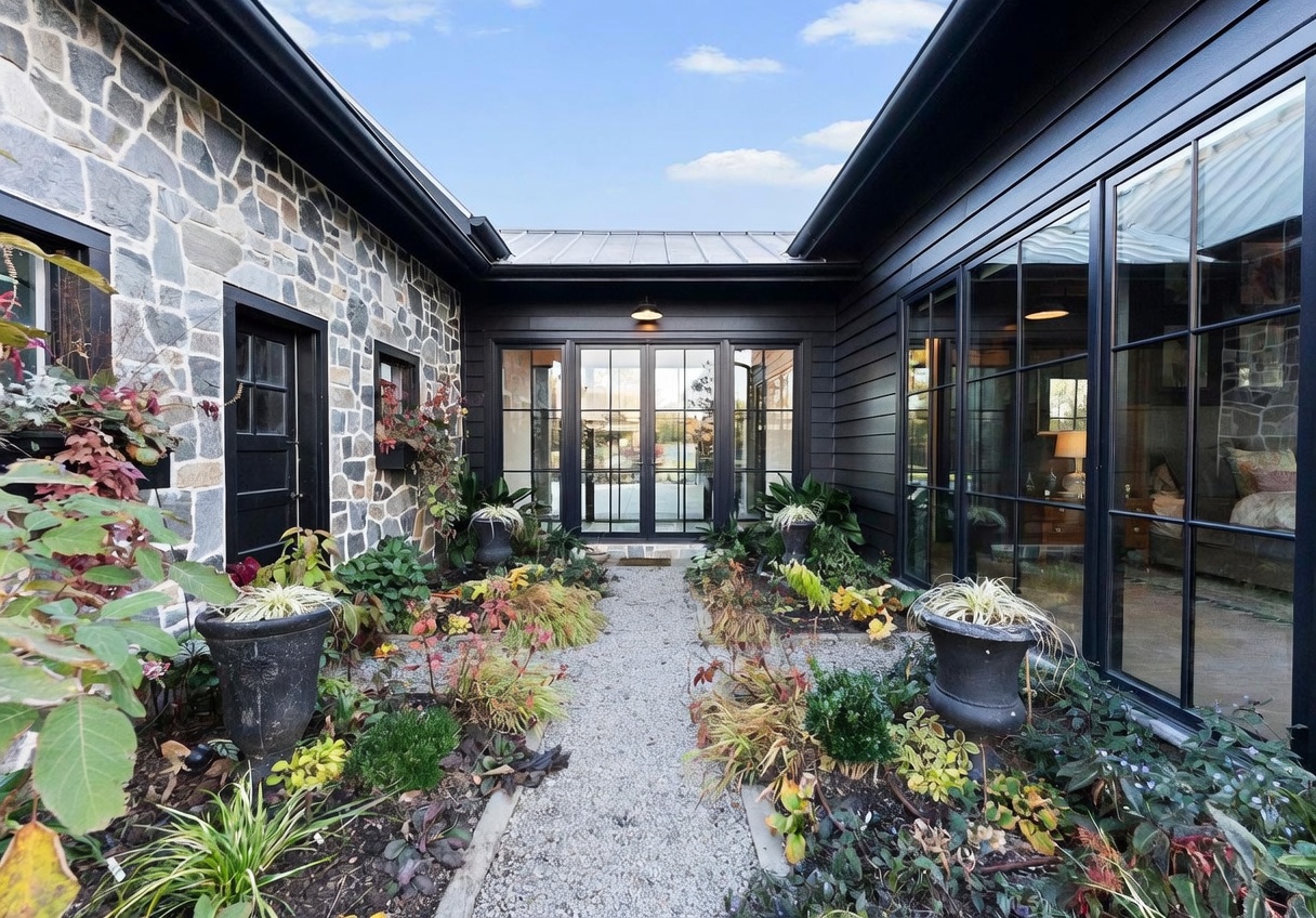 This striking courtyard entryway features a blend of rustic stone masonry on the left and modern black-paneled siding on the right. A gravel path leads through a lush garden bed filled with various shrubs and potted plants toward a set of glass-paned double doors. The perspective is centered, highlighting the architectural contrast between the traditional stone and the contemporary dark metal-framed windows that offer a glimpse into the interior living space.