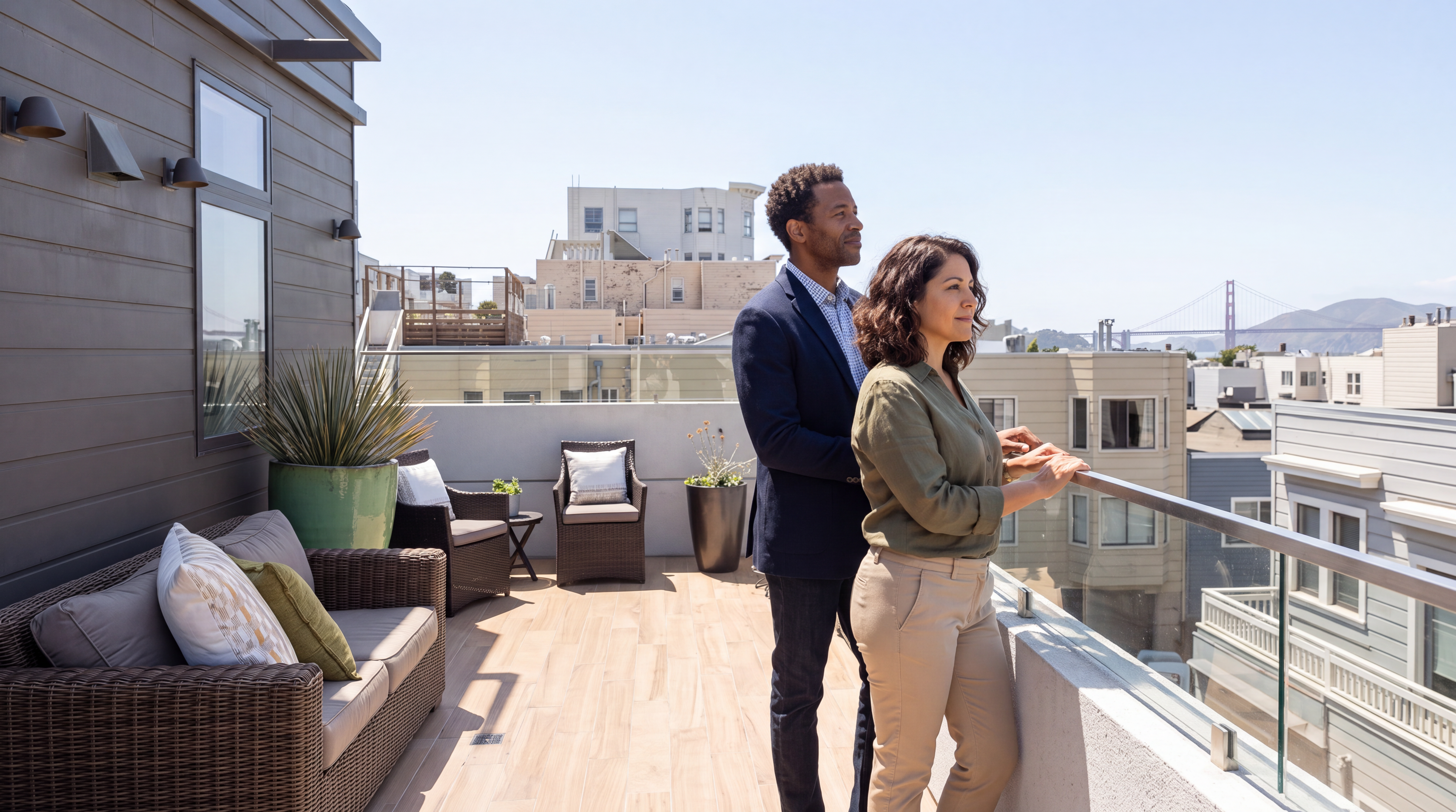 This modern, spacious rooftop balcony offers a sophisticated outdoor living area, featuring high-quality wicker lounge seating with plush cushions and contemporary tile flooring. The space is accented by elegant potted greenery and glass railings, providing an unobstructed view of the surrounding urban landscape, including the Golden Gate Bridge. It is an ideal setting for relaxation and entertainment, showcasing a refined urban lifestyle.