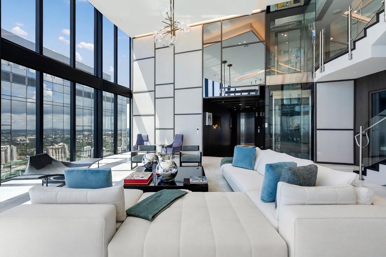 This expansive, double-height living room features dramatic floor-to-ceiling windows that offer panoramic city views and flood the space with natural light. The interior is defined by a sophisticated, modern aesthetic, showcasing a large white sectional sofa, a central coffee table with decorative accents, and a striking geometric wall treatment. A glass-enclosed elevator and a sleek staircase are visible in the background, emphasizing the high-end, architectural design of this luxury residence.
