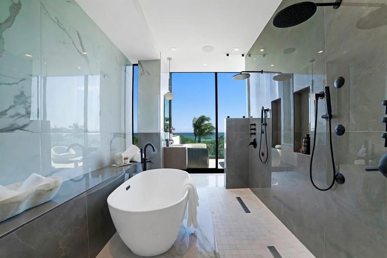 This luxurious primary bathroom features a sleek, freestanding soaking tub positioned next to a large glass-enclosed walk-in shower with multiple showerheads. The space is finished with large-format stone wall tiles and a light-colored tiled floor, creating a sophisticated, spa-like atmosphere. A wall of floor-to-ceiling glass windows provides an expansive, unobstructed view of the ocean and palm trees, bathing the room in natural light.
