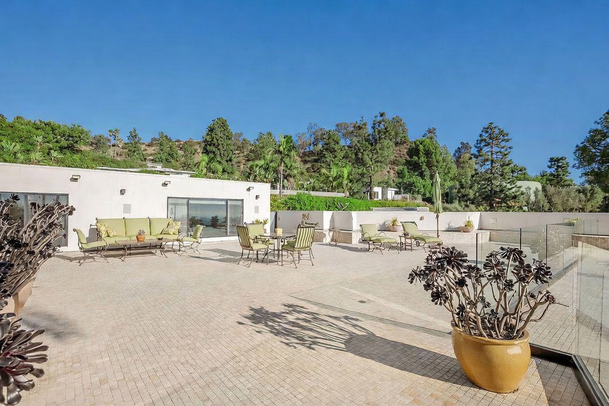 This expansive, light-toned tiled patio offers a generous outdoor living space perfect for entertaining, featuring multiple seating areas with cushioned furniture. The area is framed by a glass railing that provides an unobstructed view of the surrounding hillside landscape. The scene is captured from a slightly elevated perspective, highlighting the clean, modern aesthetic of the home's exterior and the lush greenery in the background.