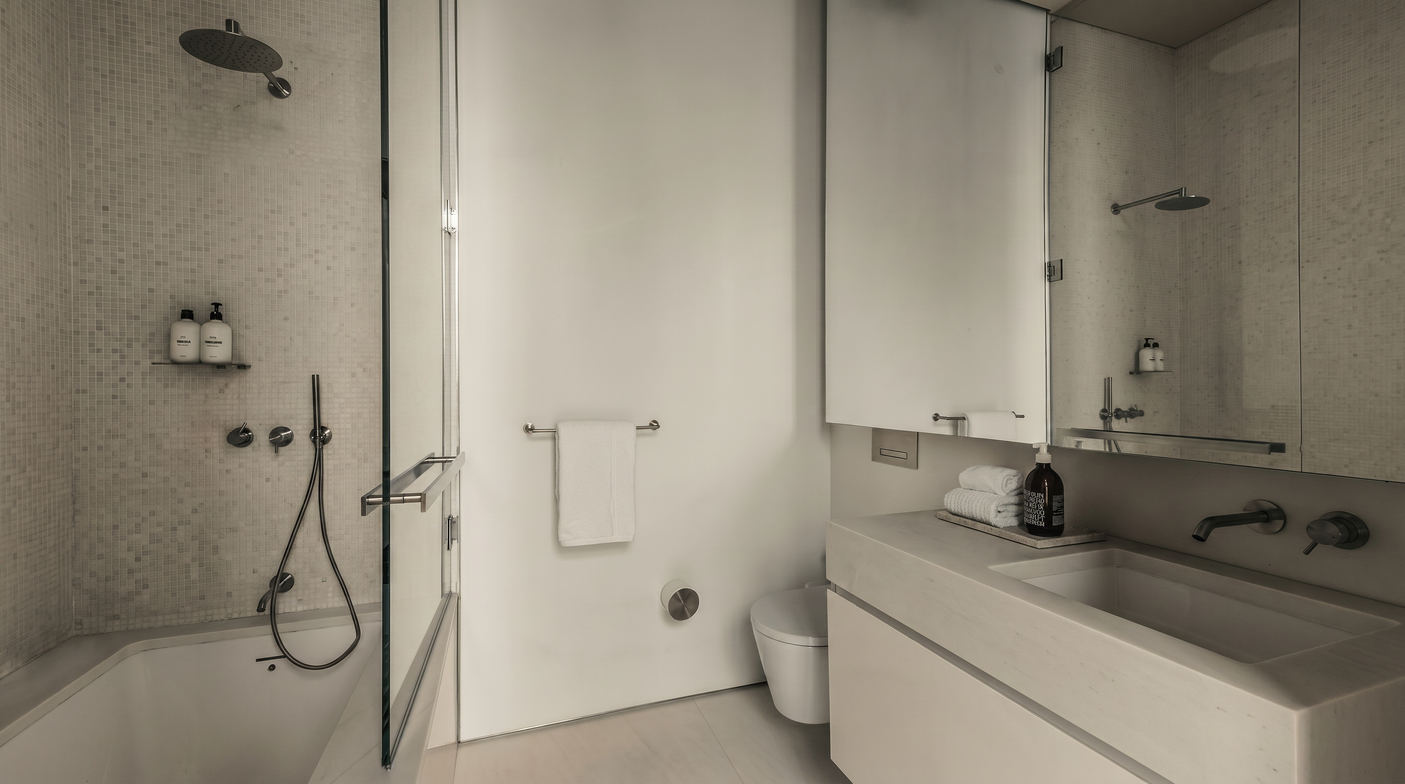 This minimalist, contemporary bathroom features a sleek, integrated vanity with a wall-mounted faucet and a large mirror that reflects the shower area. A glass-enclosed bathtub and shower combination is positioned to the left, complemented by light-toned wall textures and clean lines throughout. The space is bright and airy, emphasizing a modern aesthetic with high-end fixtures and a neutral color palette.