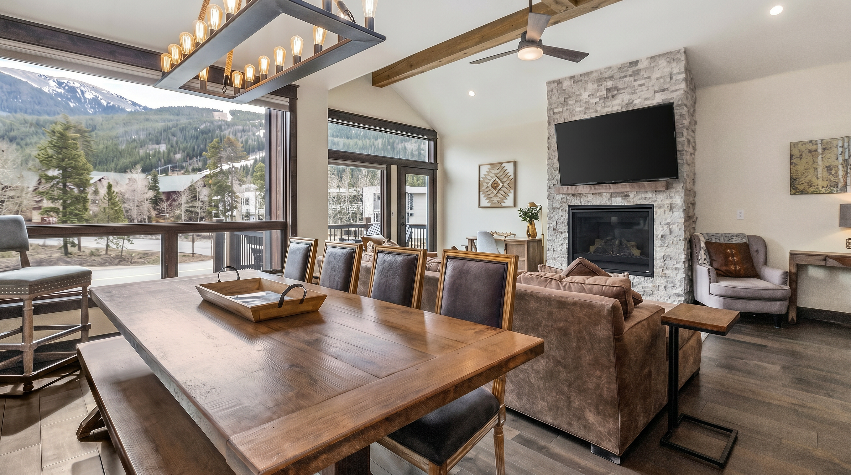 This open-concept living and dining area features a rustic mountain aesthetic with exposed wooden ceiling beams, a stone fireplace, and warm wood flooring. A large dining table sits in the foreground, while the living space is anchored by a plush brown sofa and a view of the scenic, snow-capped mountains through expansive windows. The space feels cozy and inviting, perfectly blending luxury with a natural, lodge-inspired design.