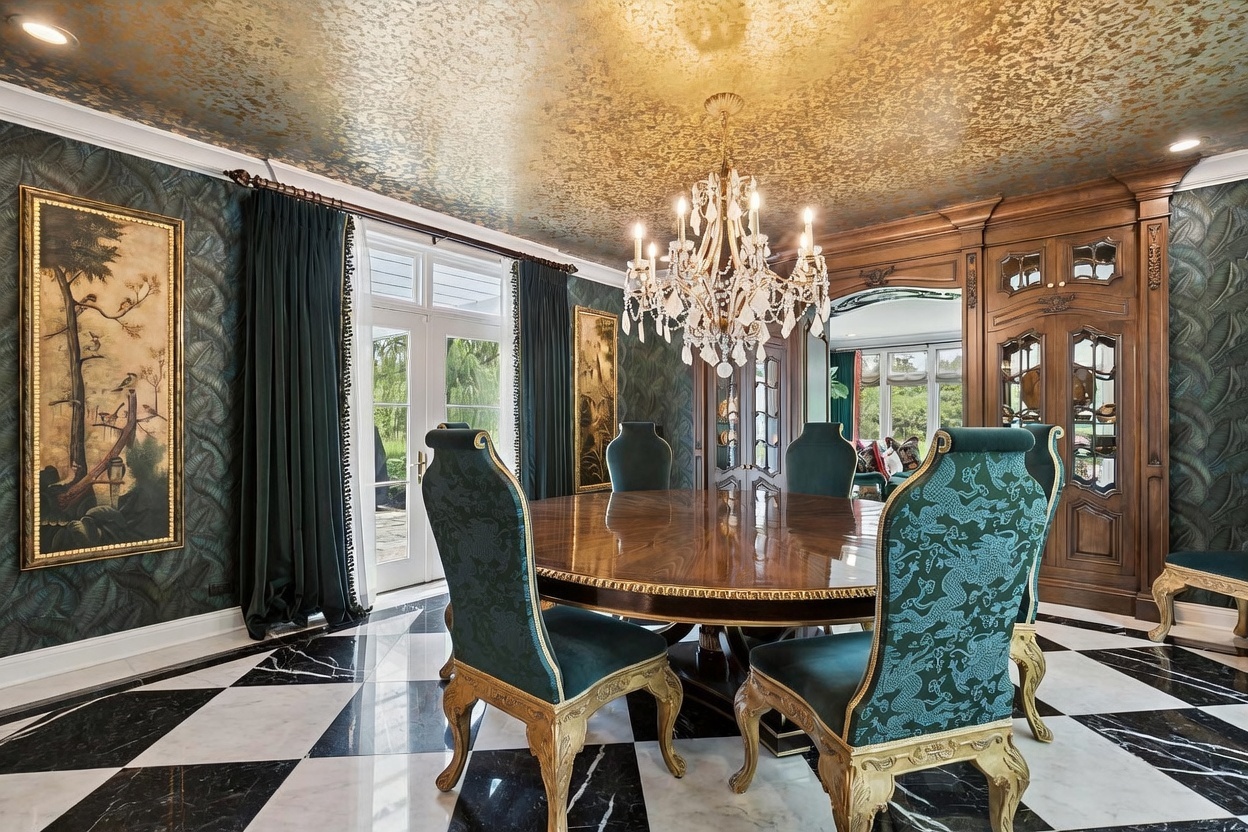 This opulent dining room features a large, round wooden table surrounded by ornate chairs upholstered in a teal dragon-patterned fabric. The space is defined by dramatic dark green patterned wallpaper, a striking gold-leaf ceiling, and a black-and-white checkered stone floor. A grand crystal chandelier hangs above the table, while a tall wooden hutch and large framed artwork add to the sophisticated, formal aesthetic.