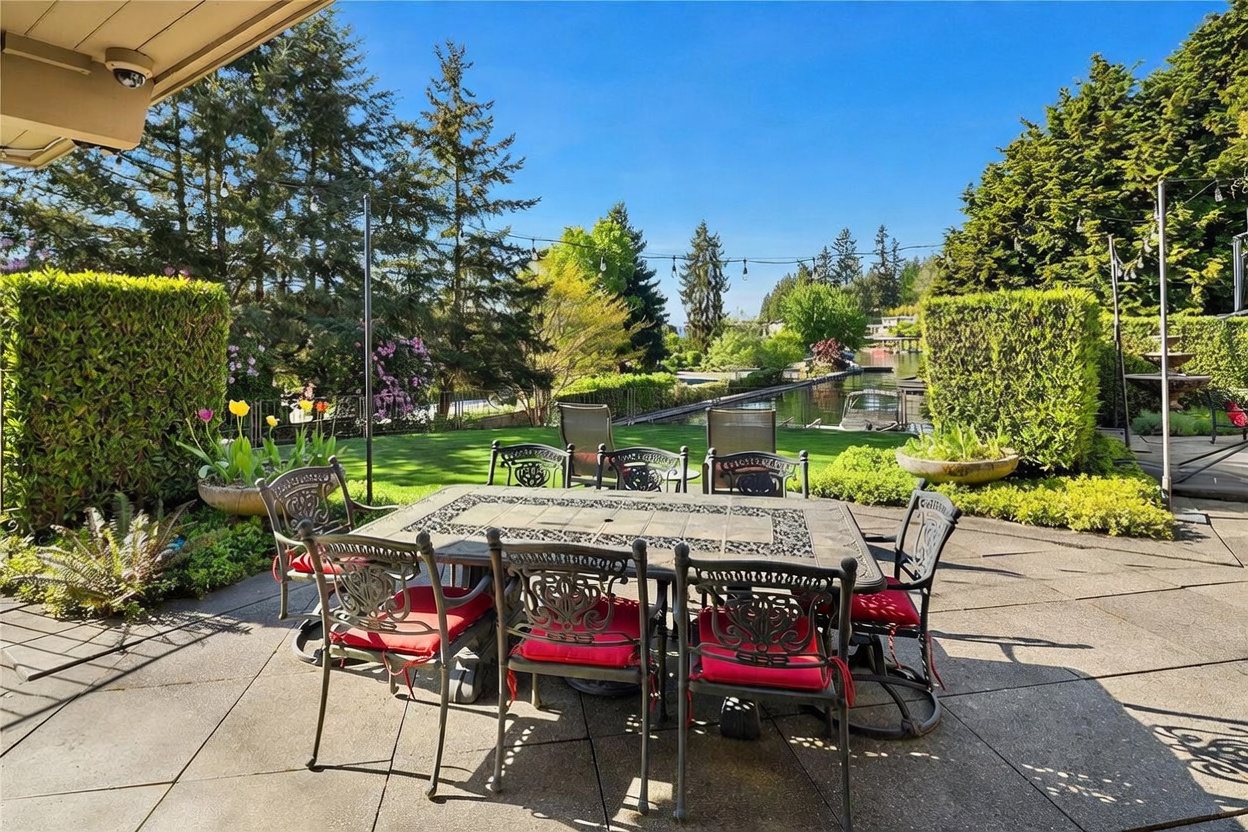 This outdoor patio space features a large metal dining set with red cushions, positioned on a paved surface overlooking a lush green lawn and a private canal. The scene is framed by manicured hedges and mature trees, with string lights hanging above to create an inviting atmosphere for al fresco dining. The perspective is from the patio looking out toward the water, capturing a serene and well-maintained backyard setting.