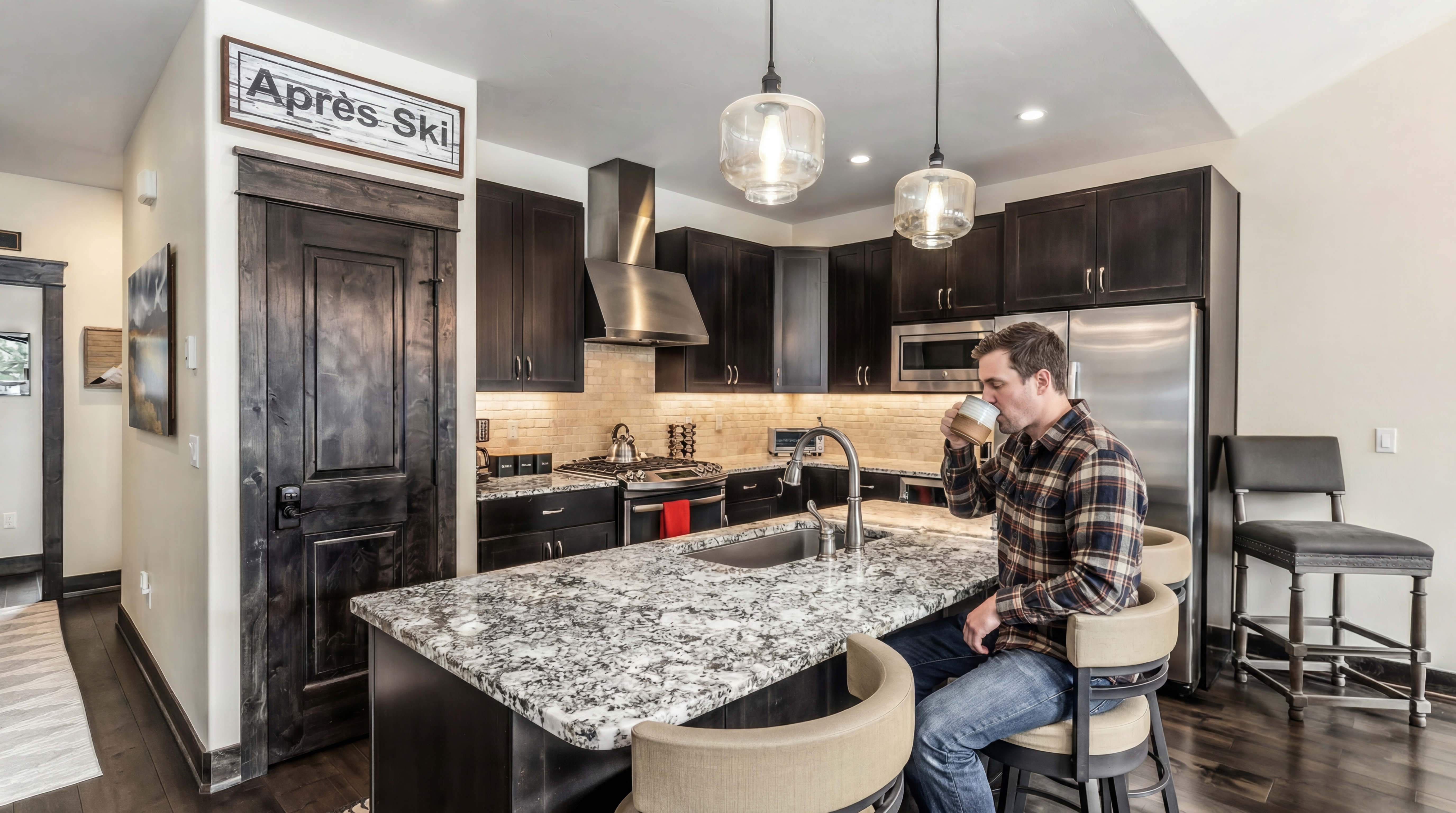 This modern kitchen features dark wood cabinetry, stainless steel appliances, and a striking granite island with seating for four. The space is accented by warm pendant lighting, a tiled backsplash, and an 'Après Ski' sign above the rustic wooden door, creating a cozy, mountain-inspired atmosphere. The perspective captures the functional layout of the kitchen, highlighting the seamless flow between the cooking area and the breakfast bar.