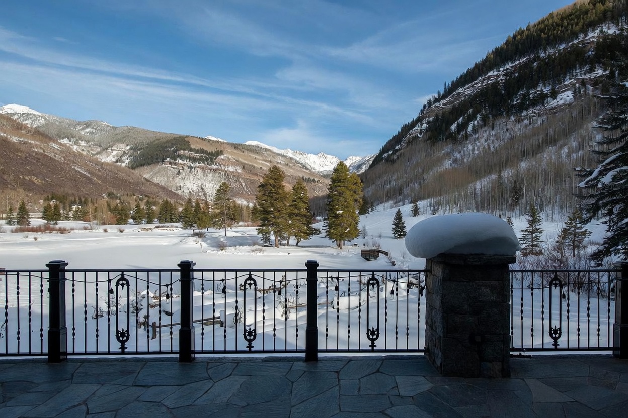 This image captures a stone-paved patio featuring a decorative black metal railing that overlooks a vast, snow-covered mountain landscape. The perspective is from a slightly elevated vantage point, showcasing a serene winter scene with tall evergreen trees and distant, rugged peaks under a clear blue sky. A stone pillar topped with a thick layer of snow anchors the foreground, emphasizing the cold, tranquil atmosphere of this mountain property.