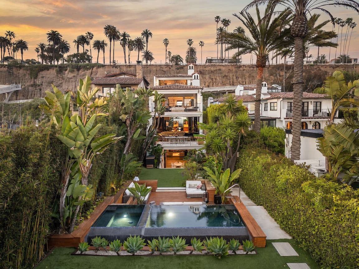 This rear view of a multi-story coastal residence showcases a tiered architectural design with expansive balconies and large glass doors opening to the interior living spaces. The property features a private backyard with a modern rectangular pool and integrated spa, surrounded by lush tropical landscaping and tall bamboo hedges. The scene is captured at sunset, highlighting the warm glow of the home's interior lighting against the dramatic cliffside backdrop.