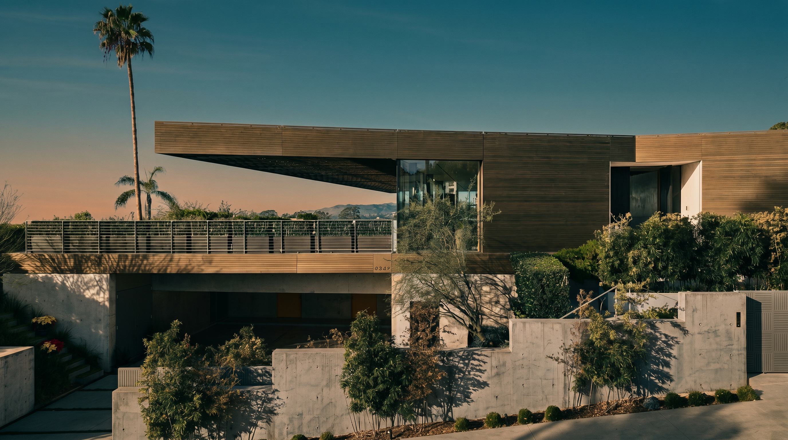 This striking modern residence features a bold, cantilevered roofline and a sophisticated mix of warm wood-toned siding and raw concrete elements. The multi-level design includes a spacious elevated terrace with a sleek metal railing, overlooking a lower-level garage area and landscaped grounds. The architectural style is contemporary and minimalist, emphasizing clean lines and a seamless integration with the surrounding environment.