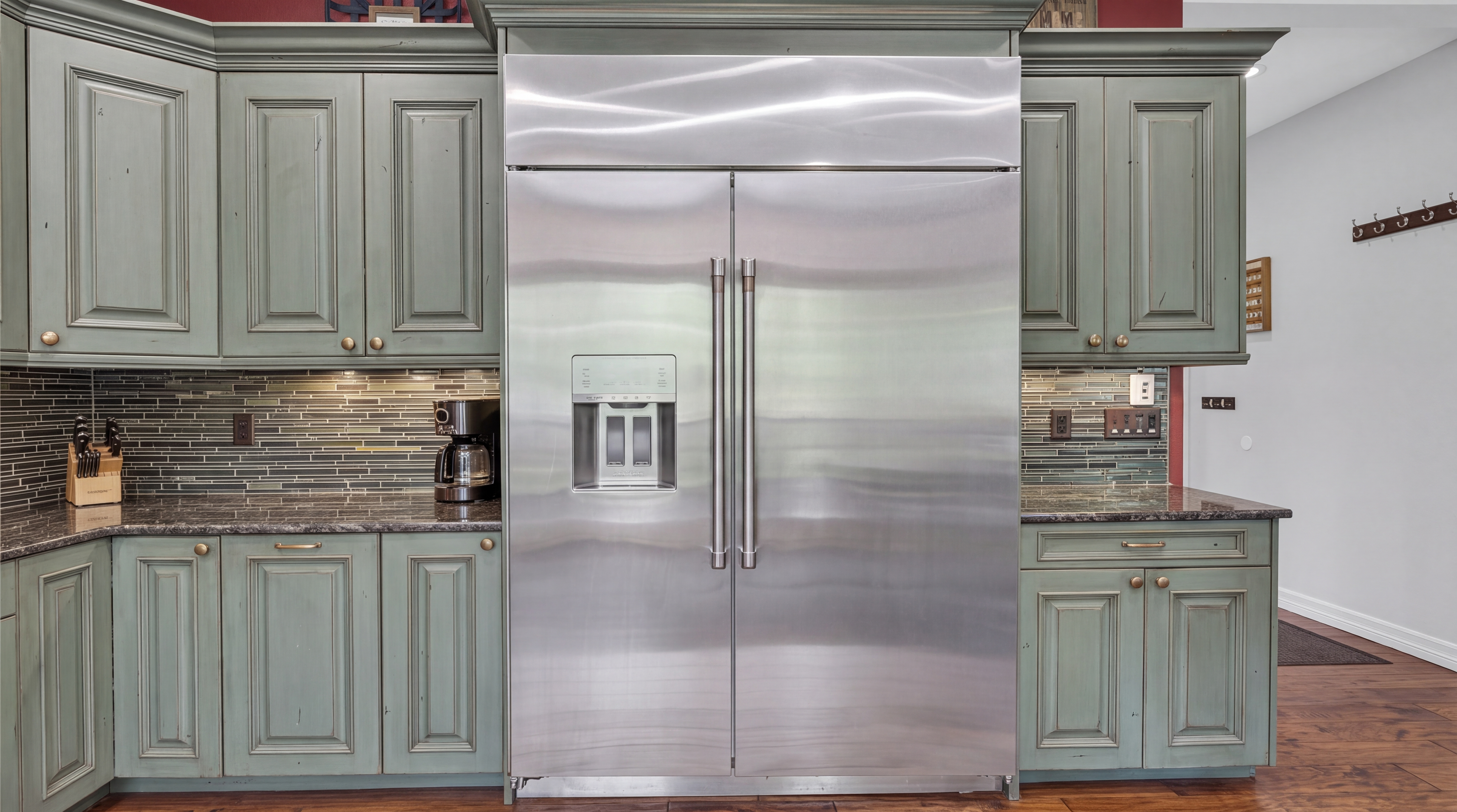 This kitchen features custom green-hued distressed cabinetry paired with dark granite countertops and a contemporary glass tile backsplash. A large, side-by-side stainless steel refrigerator with an integrated water dispenser serves as the focal point of the wall, flanked by ample storage space. The aesthetic blends a rustic, traditional warmth with functional, modern appliance selections.