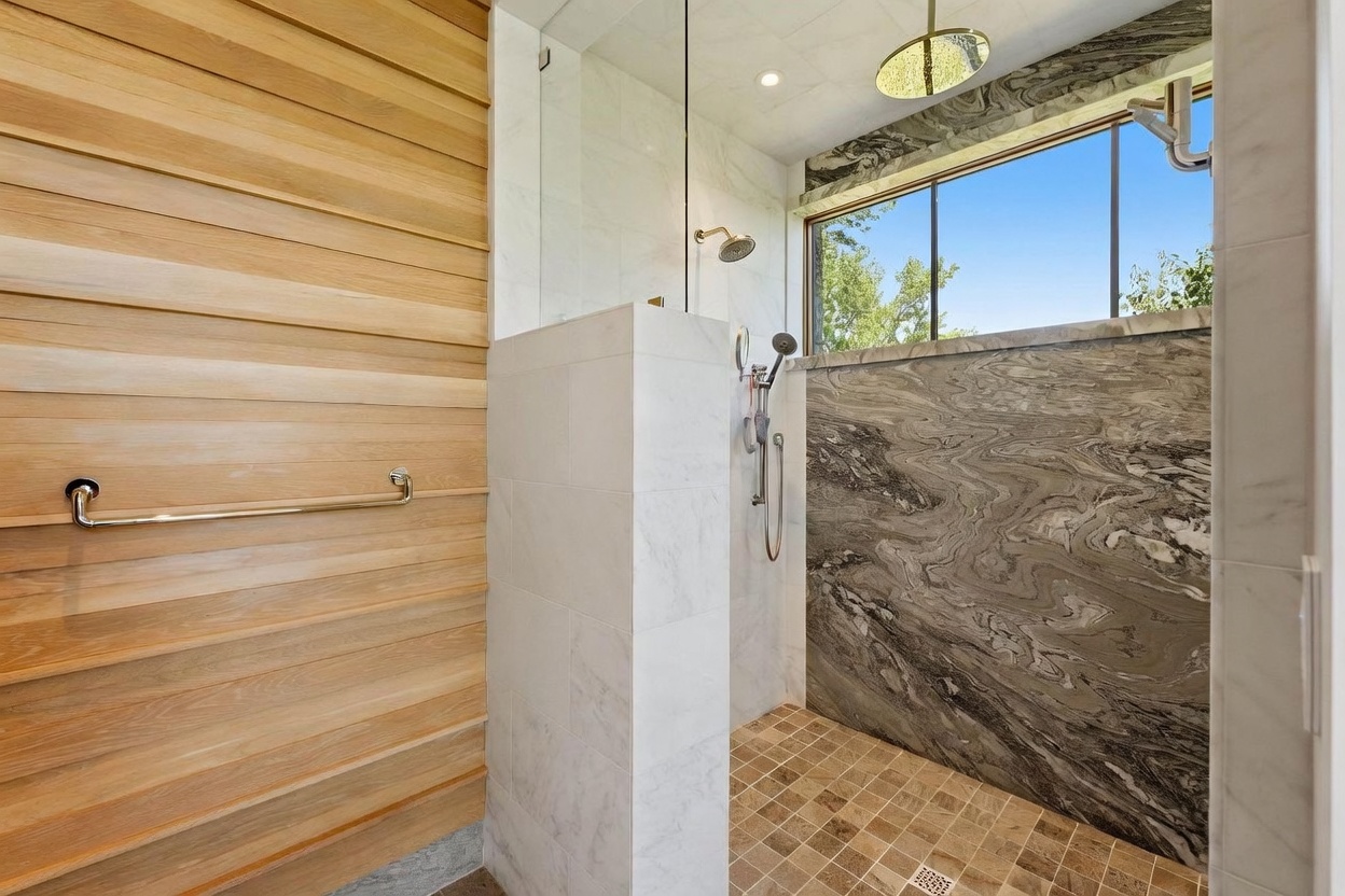 This modern walk-in shower features a striking contrast between light wood-paneled walls and a dramatic, dark-veined stone accent wall. The space includes a glass-enclosed shower area with both a rainfall showerhead and a handheld sprayer, complemented by a window that brings in natural light. The floor is finished with small, square-patterned tiles, creating a sophisticated and spa-like atmosphere.