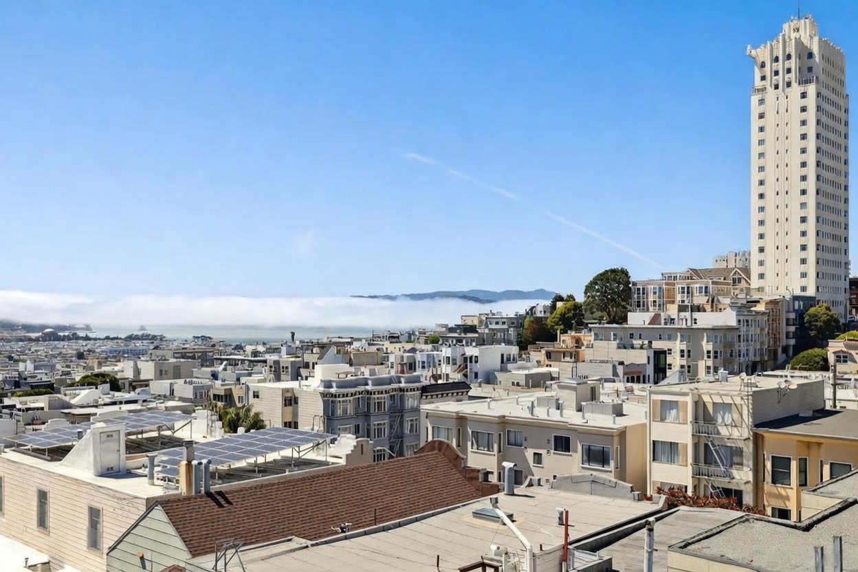 This elevated aerial perspective captures a dense urban cityscape under a clear blue sky, featuring a prominent high-rise tower as a focal point. The foreground showcases various rooftops, some equipped with solar panels, while the background reveals a layer of low-hanging fog rolling over the distant bay. The scene provides a comprehensive view of the neighborhood's architectural density and its proximity to the coastal landscape.