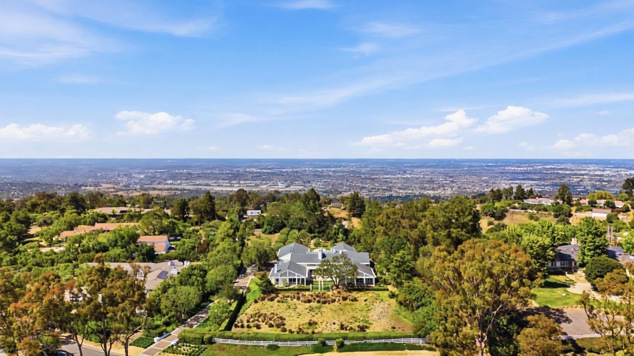 This high-angle aerial view captures a sprawling estate nestled within a lush, tree-filled landscape overlooking a vast valley and distant city skyline. The primary residence features a classic architectural style with a multi-gabled roofline, surrounded by manicured lawns, mature trees, and winding pathways. The perspective emphasizes the property's expansive grounds and its elevated, scenic position.