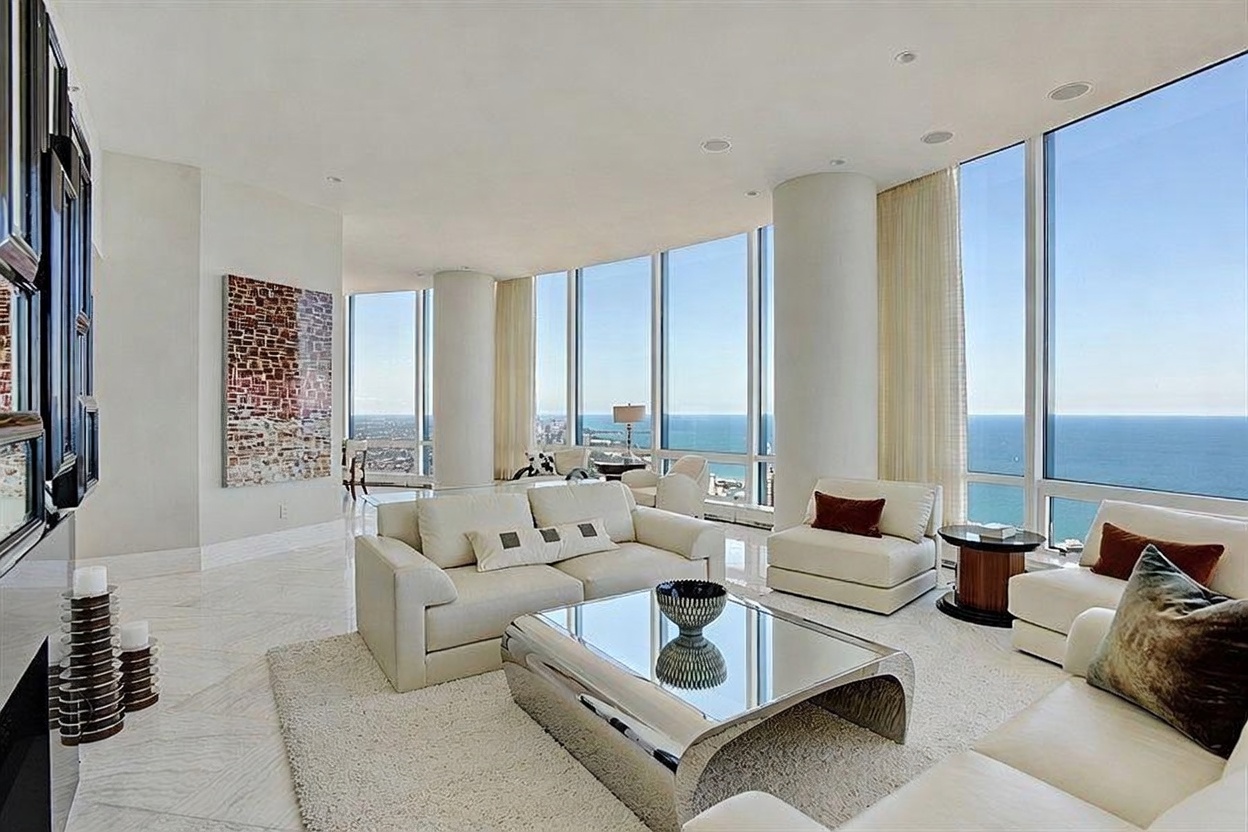 This expansive, high-rise living room features a sophisticated, modern aesthetic with light-toned stone flooring and a neutral color palette. The space is defined by floor-to-ceiling windows that provide panoramic views of the lake and city skyline, flooding the room with natural light. Furnishings include a plush white sofa, matching armchairs, and a reflective, metallic-finished coffee table, all arranged on a soft area rug to create a luxurious and airy atmosphere.