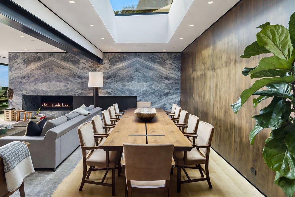 This sophisticated open-concept dining area features a long, light wood table surrounded by twelve upholstered chairs, positioned beneath a large skylight that floods the space with natural light. To the left, a comfortable living area includes a sofa and a linear fireplace set into a dramatic, dark-veined stone wall. The room is anchored by a warm wood-paneled wall on the right and a large potted plant, creating a luxurious and inviting atmosphere.