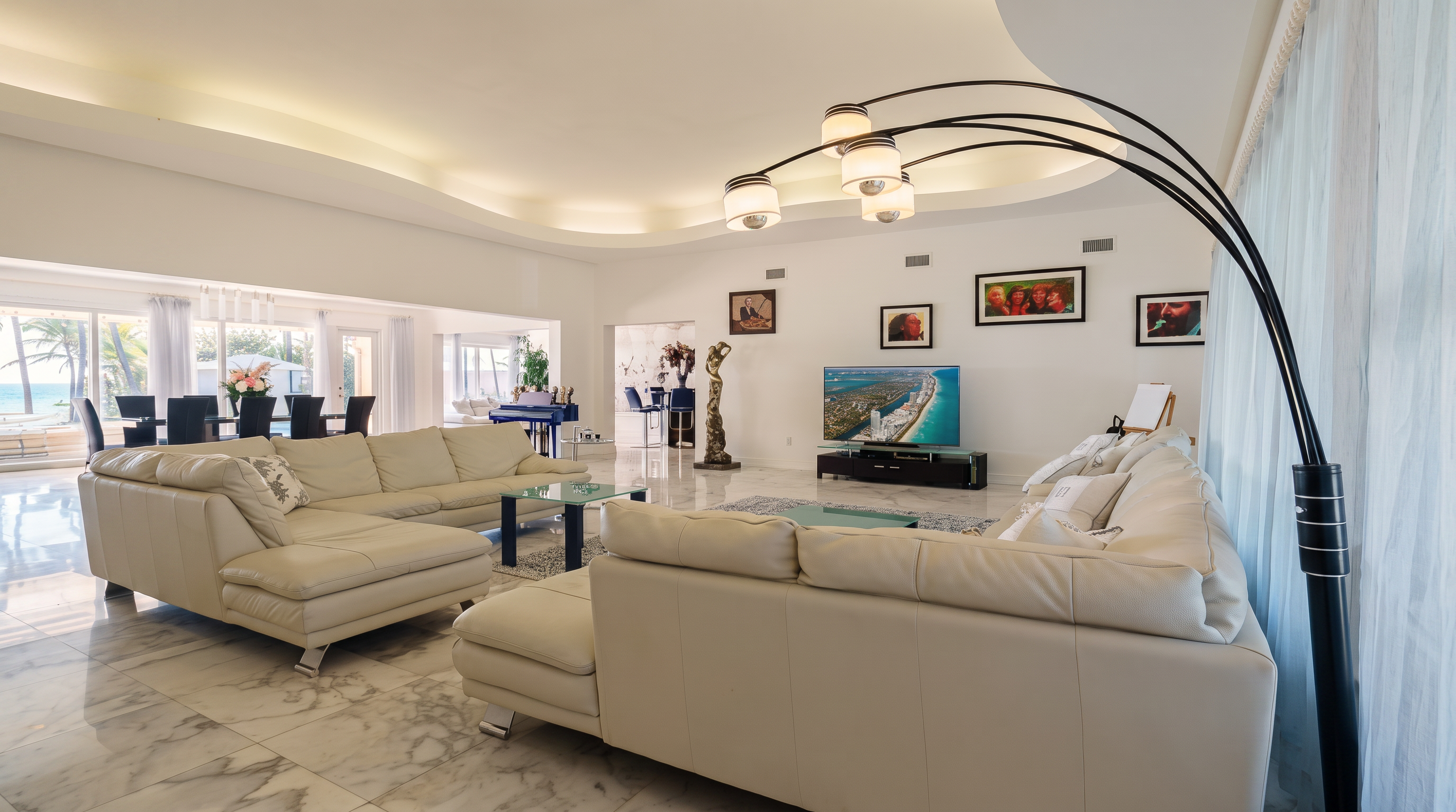 This spacious, open-concept living area features light-colored, large-format stone flooring and a curved, recessed ceiling design with integrated lighting. The room is furnished with two large, cream-colored sectional sofas arranged around a glass-topped coffee table, with a television mounted on the wall as a focal point. A tall, arched floor lamp curves over the seating area, and the space flows seamlessly into a dining area visible through a wide opening in the background.