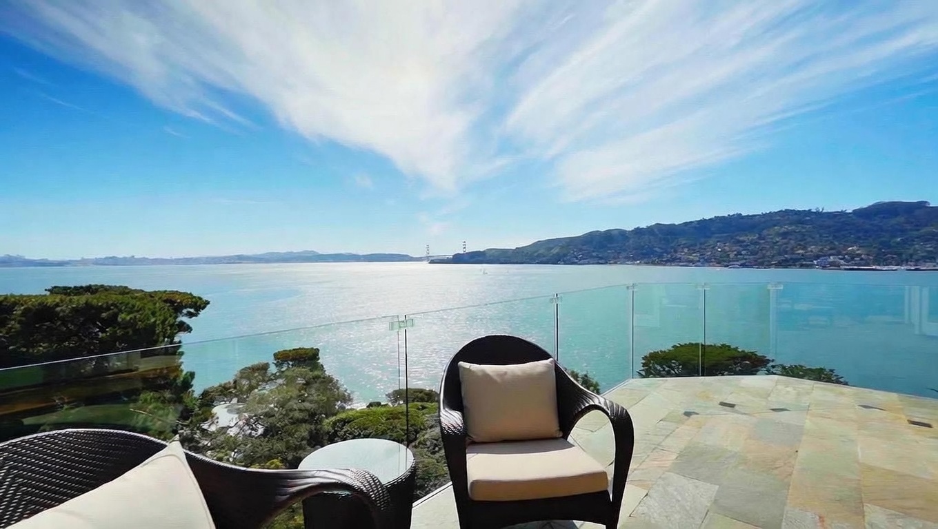 This elevated balcony offers a panoramic, unobstructed view of a large bay with distant hills and a bridge visible on the horizon. The space is furnished with dark wicker chairs featuring light-colored cushions and a small side table, set upon a stone-tiled floor. A sleek, frameless glass railing ensures the scenery remains the focal point, creating an open and airy atmosphere perfect for relaxation.
