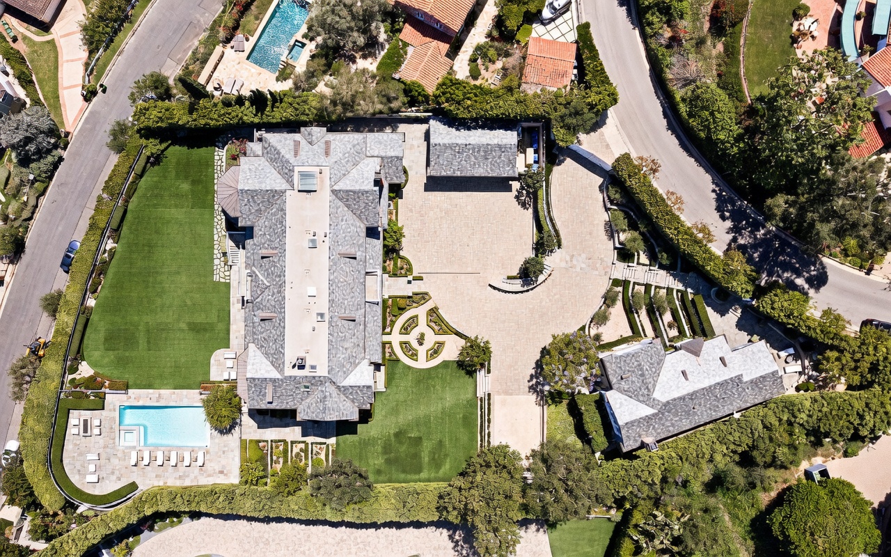 This high-angle aerial view captures a sprawling luxury estate featuring a large main residence with a complex, multi-gabled roofline. The property includes a manicured lawn, a rectangular swimming pool with an adjacent patio area, and a secondary structure, all surrounded by dense, mature landscaping. A paved circular driveway leads to a detached garage, showcasing the expansive and private nature of the grounds.
