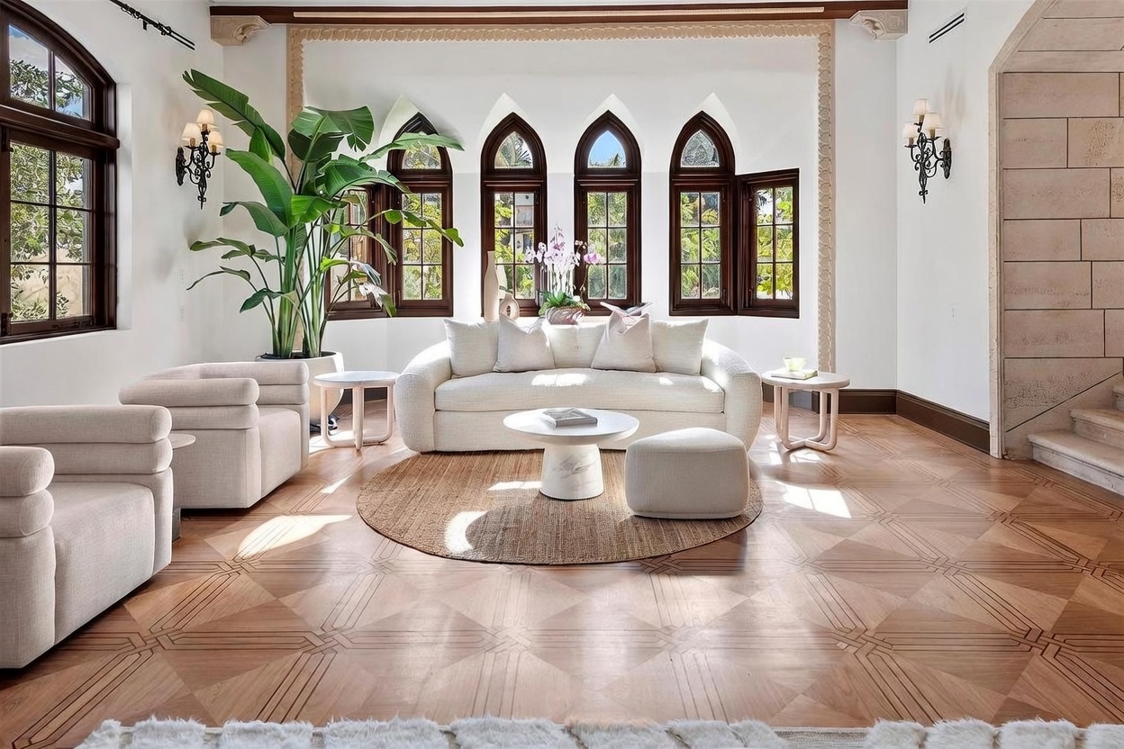This elegant living room features a sophisticated neutral color palette with a plush white sofa, matching armchairs, and a circular coffee table centered on a textured rug. The space is defined by its striking architectural details, including a series of arched, dark-framed windows and intricate patterned wood flooring. A large potted plant adds a touch of greenery, while wall-mounted sconces provide soft lighting, creating a bright and airy atmosphere.