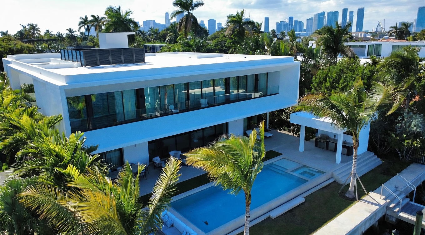 This aerial perspective captures a modern, two-story luxury residence featuring a flat roof and extensive glass walls that offer a seamless indoor-outdoor connection. The property includes a rectangular swimming pool set within a landscaped yard, complemented by a covered patio area and lush tropical palm trees. In the background, a panoramic view of a city skyline across the water provides a sophisticated urban backdrop to this serene waterfront estate.