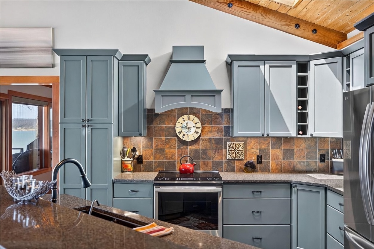 This kitchen features blue-toned cabinetry paired with a dark stone countertop and a rustic, multi-colored tile backsplash. A central stove with a matching range hood serves as the focal point, accented by a decorative wall clock. The space is framed by a vaulted ceiling with exposed wood beams and offers a glimpse of a scenic outdoor view through an adjacent doorway.
