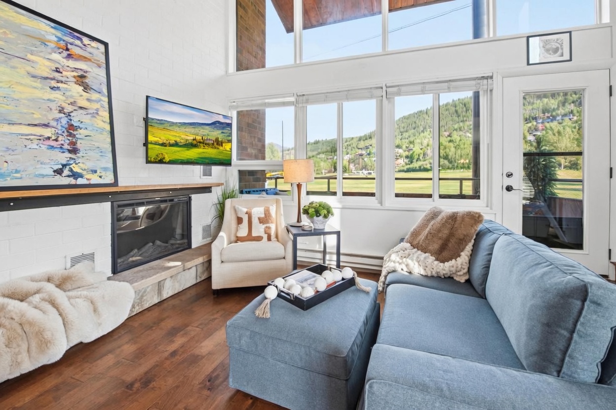 This bright and inviting living room features a cozy blue sofa with a matching ottoman, a comfortable armchair, and a fireplace with a stone hearth. Large windows provide an expansive view of the surrounding mountains and greenery, filling the space with natural light. The room is decorated with a large abstract painting, a textured throw blanket, and a tray with decorative spheres, creating a warm and relaxing atmosphere.
