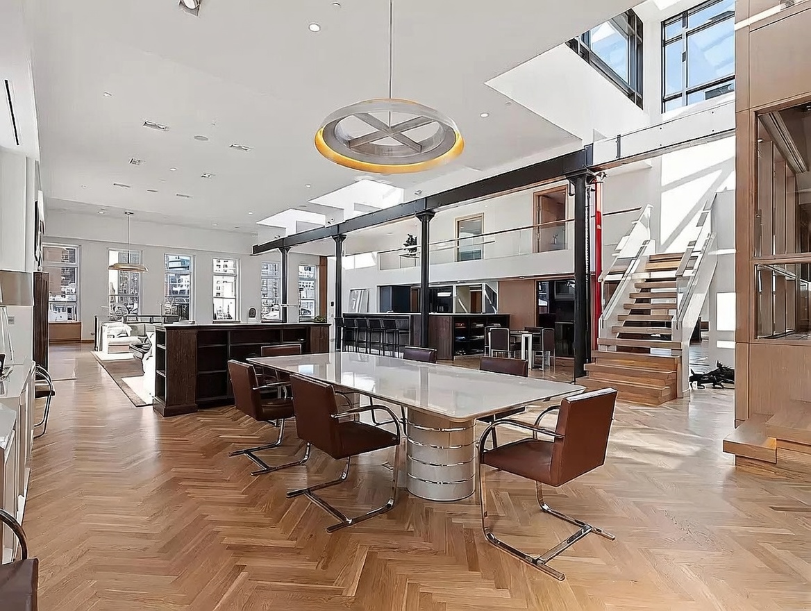 This expansive, open-concept living and dining area features a high-ceilinged industrial aesthetic with exposed structural columns and a prominent circular pendant light. A large dining table with leather chairs sits on a herringbone-patterned wood floor, while a staircase with glass railings leads to an upper mezzanine level. The space is flooded with natural light from large windows and skylights, creating an airy and sophisticated atmosphere.
