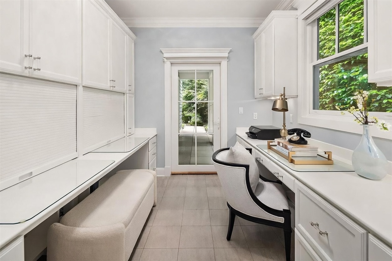 This bright and organized home office features dual built-in desk stations with white cabinetry and glass-topped surfaces. A central glass-paned door provides a view into a lush, green outdoor space, while a comfortable upholstered bench and a stylish desk chair complete the workspace. The room is finished with light-toned walls and neutral tile flooring, creating a clean and professional atmosphere.