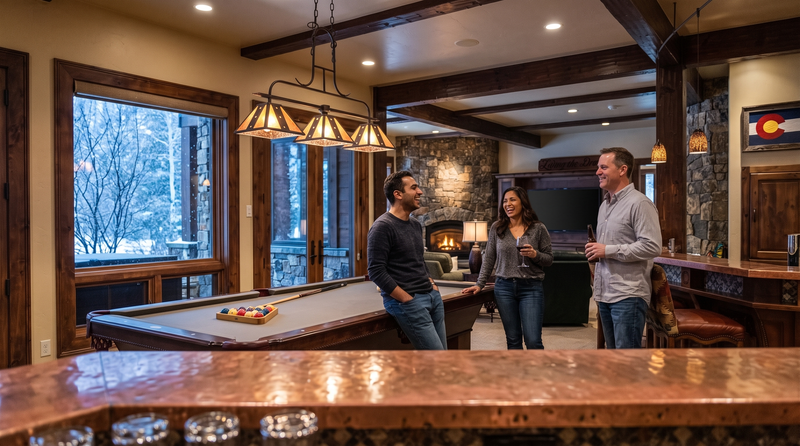 This spacious, lodge-style basement serves as an ideal entertainment area, featuring a pool table under custom pendant lighting and a wet bar with a rich copper countertop. The room incorporates warm elements like a stone fireplace, wood beams, and large windows with snowy exterior views, creating a cozy and inviting atmosphere for social gatherings.