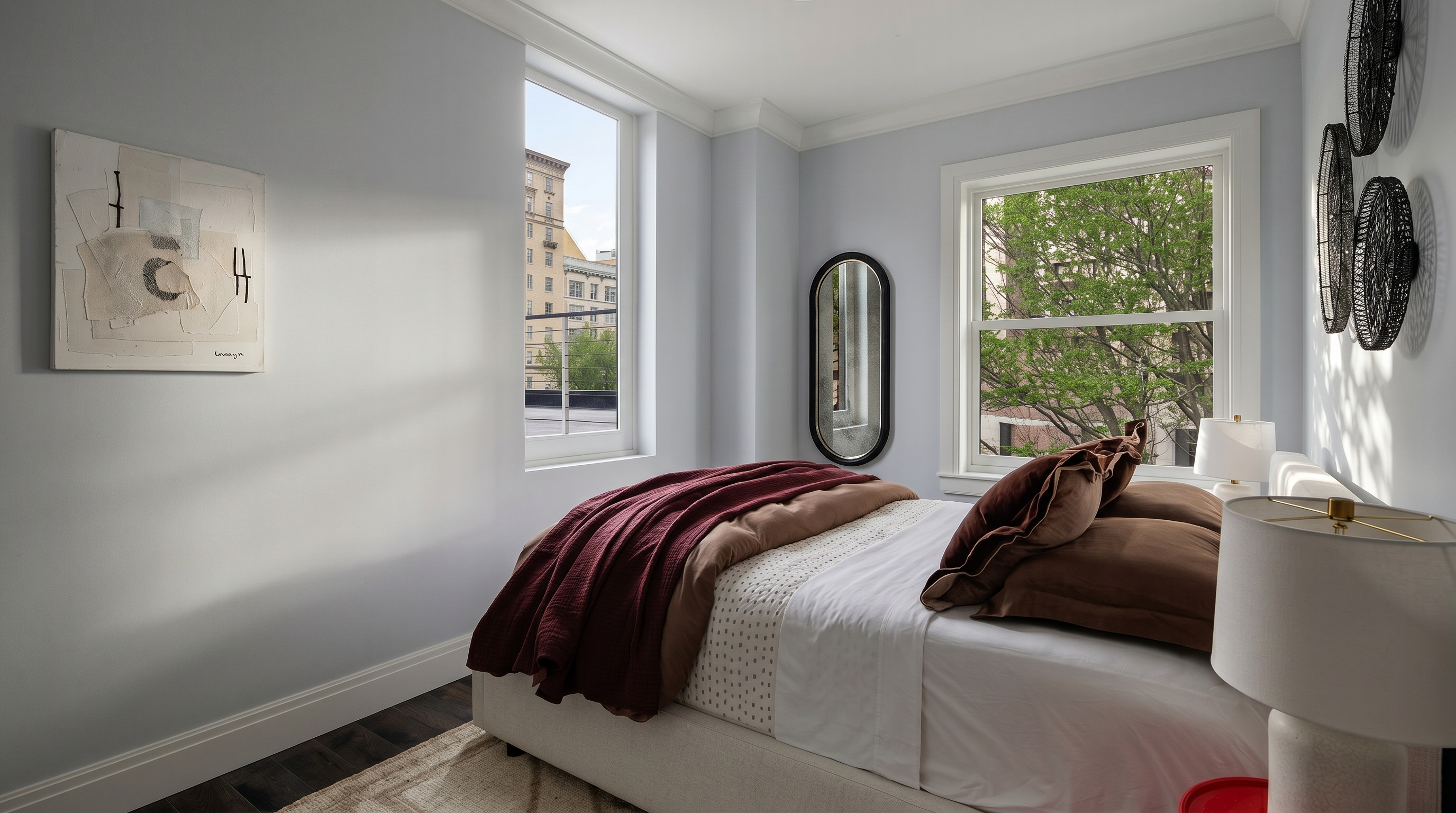 This cozy and well-lit bedroom features a neutral color palette with light blue walls and dark hardwood floors, creating a serene atmosphere. The room is furnished with a plush bed dressed in layered bedding, a modern bedside lamp on a vibrant red side table, and a minimalist oval mirror. Large windows allow for ample natural light and offer a pleasant view of the surrounding neighborhood trees and buildings.