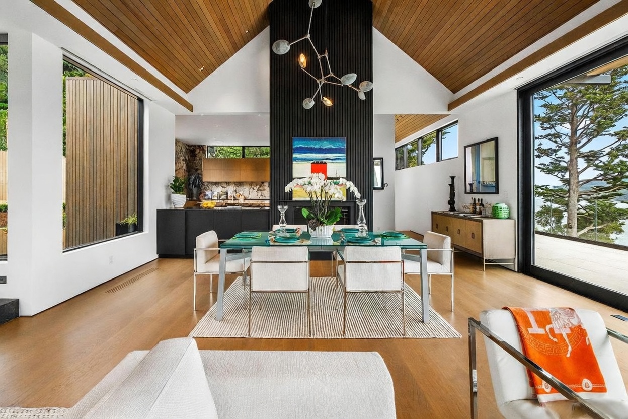 This bright, open-concept dining area features a glass-topped table centered beneath a modern, multi-armed chandelier. The space is defined by a tall, dark-paneled chimney breast that anchors the room, while light wood flooring and a vaulted wood-paneled ceiling create a warm, airy atmosphere. Large glass sliding doors on the right provide a seamless connection to the outdoors, offering expansive views of the surrounding trees and water.