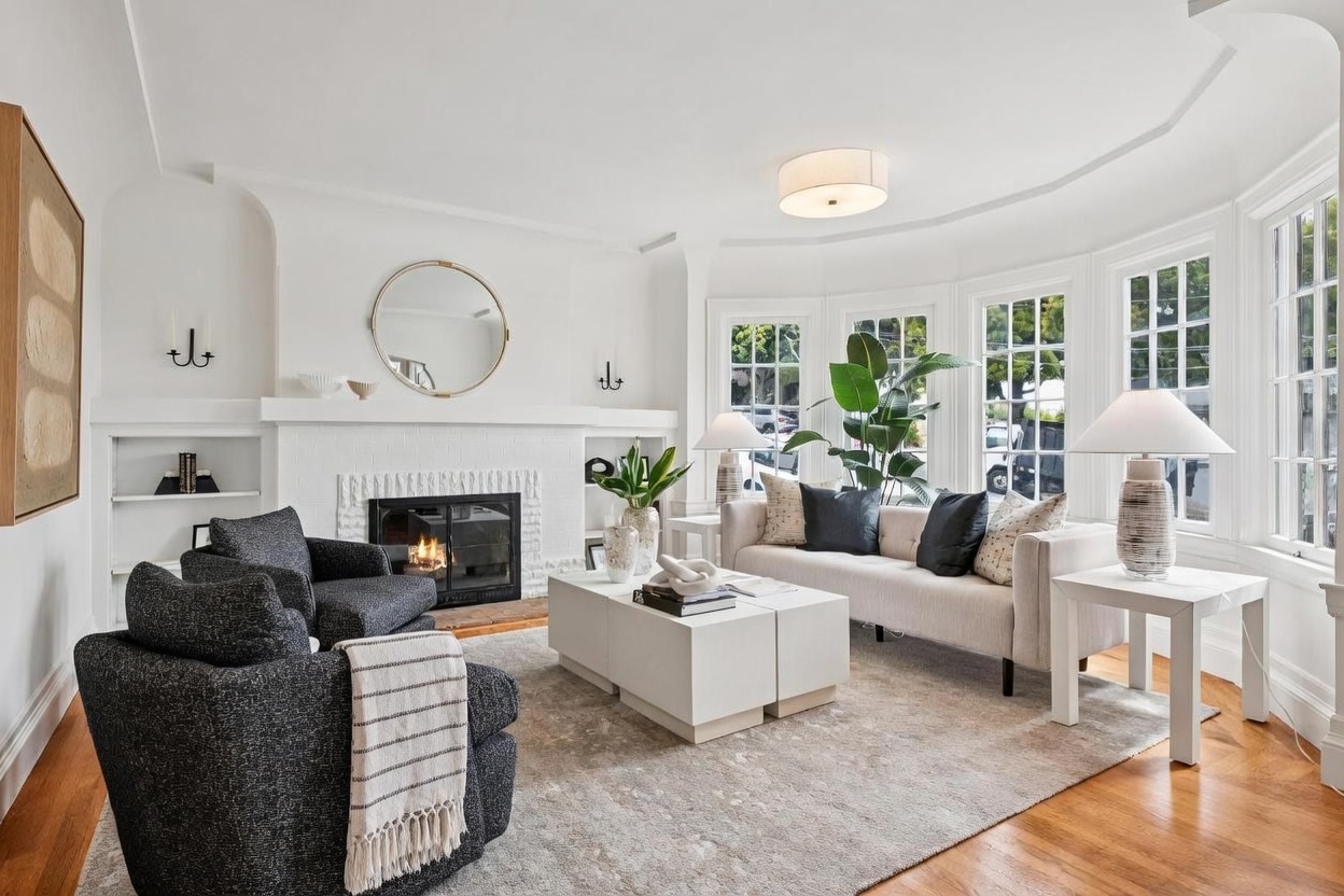 This bright and airy living room features a white-painted brick fireplace as a central focal point, complemented by a large round mirror and built-in shelving. The space is furnished with a neutral-toned sofa and two dark textured armchairs arranged around a pair of white coffee tables on a large area rug. Large bay windows allow for ample natural light, creating an inviting and sophisticated atmosphere.